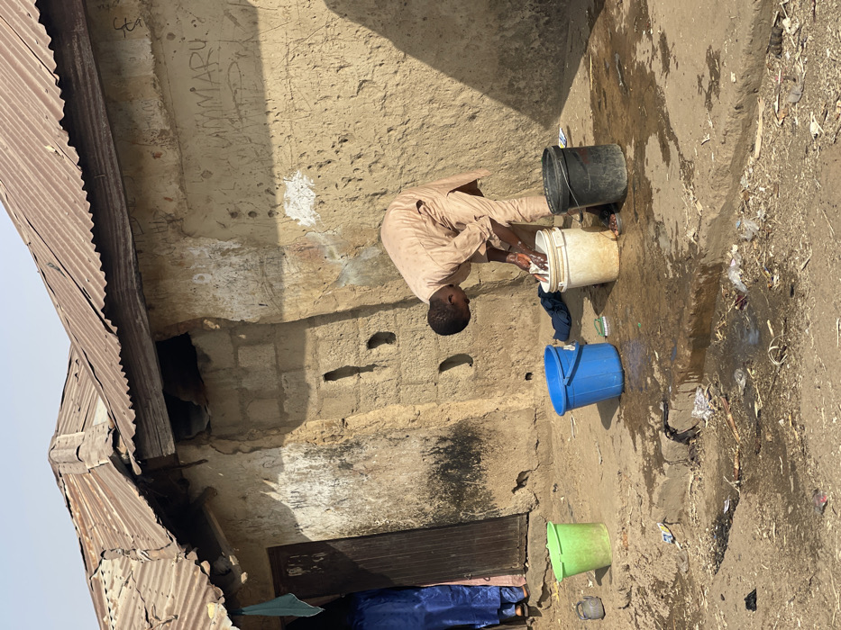 The scene depicts an outdoor activity in Unguwar Gabas, Nigeria, occurring under clear daylight. A male individual, dressed in a light-colored tan or beige tunic, is bent over, actively engaged in hand-washing items within a white plastic bucket. His hands are submerged in the water inside the bucket. Surrounding him on the uneven, dirt ground are two other plastic buckets: one blue, partially filled with liquid, and one black. A green plastic bucket and a small dark pot are positioned further left, near a wooden doorway. A blue fabric item and a small scrub brush are visible on the ground near the blue bucket. Water spillage from the washing activity is evident on the dirt and concrete ground, mixing with scattered debris, including plastic bags and wrappers.
The background consists of a weathered building with walls constructed from mud bricks and possibly some concrete blocks, exhibiting discoloration and erosion. The word "UMMARU" is visible as graffiti etched into the upper wall. The building is topped with a rusty, corrugated metal roof. A second individual, wearing a blue garment, is partially visible standing inside the doorway to the left, observing the scene.