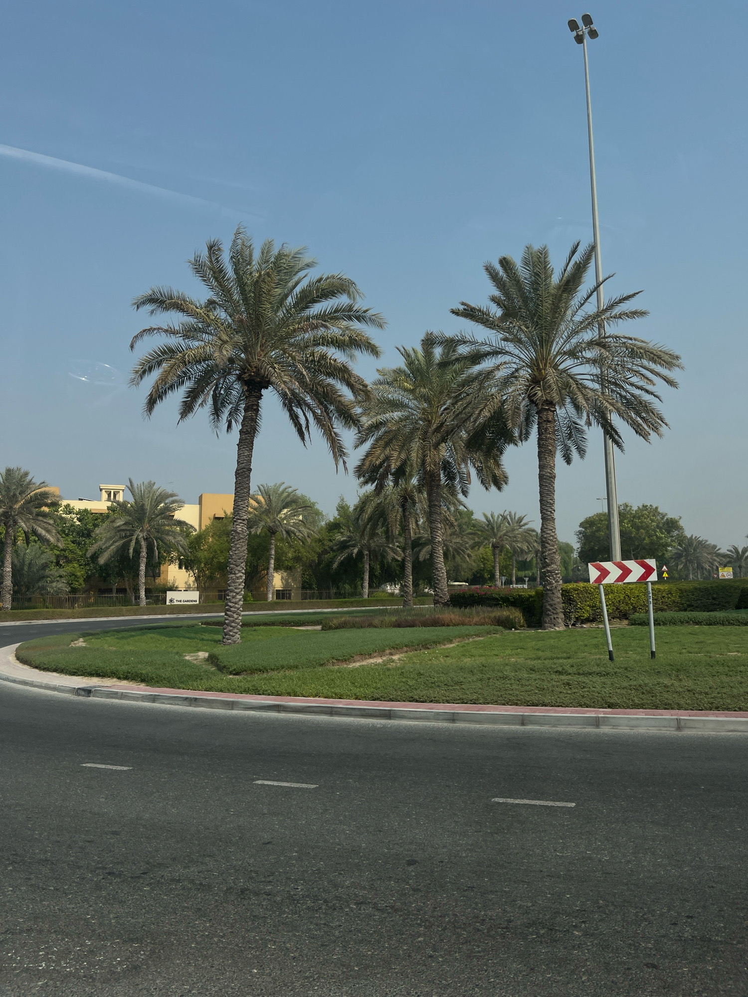 The image shows a view of a street with palm trees and a building in the background. The street is paved with asphalt and has white lane markings. There is a circular grassy median with several palm trees. A tall street light with multiple lamps stands on the right side of the image, and a traffic sign indicating a curve to the right is also visible. In the background, there is a low-rise building with a light yellow facade and some greenery. The sky is clear and blue, with a faint contrail visible in the upper left corner. The environment appears to be a well-maintained urban or suburban area in Dubai, United Arab Emirates, possibly in a residential or commercial district like "The Gardens," as suggested by a sign in the background. No people or vehicles are present in the frame.