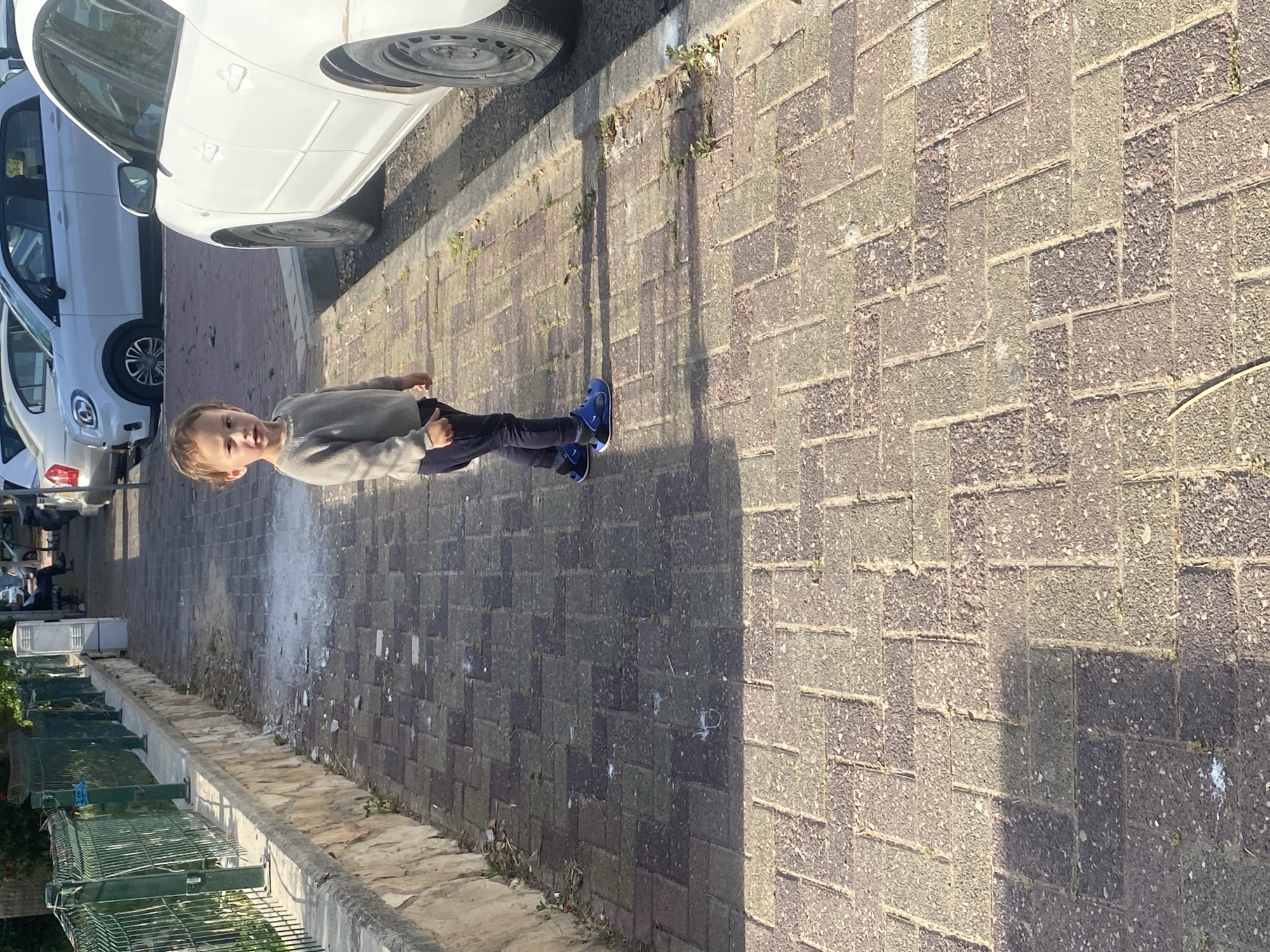 A child stands on a brick sidewalk in Kiryat Bialik, Israel. The child is wearing a gray sweater, dark pants, and blue sneakers. To the left are parked cars and a green fence. The sidewalk is composed of brown and gray bricks. The scene is lit with sunlight, casting shadows.