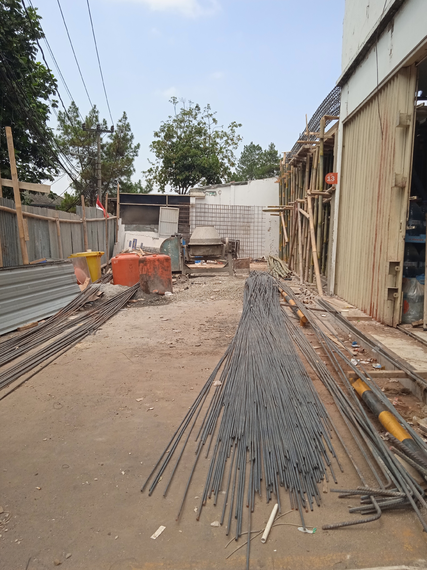 The image depicts a construction site in Cileunyi, Indonesia. In the foreground, a large pile of rebar rods is neatly arranged on the dirt ground. To the left, more rebar rods are laid out, along with corrugated metal sheeting and wooden scaffolding. In the middle ground, two large orange water tanks and a yellow plastic bin are visible, positioned near a concrete mixer and other construction equipment. The background reveals trees, utility poles with wires, and a white building with a corrugated metal facade. The sky is overcast with scattered clouds. There are no people visible in the scene.