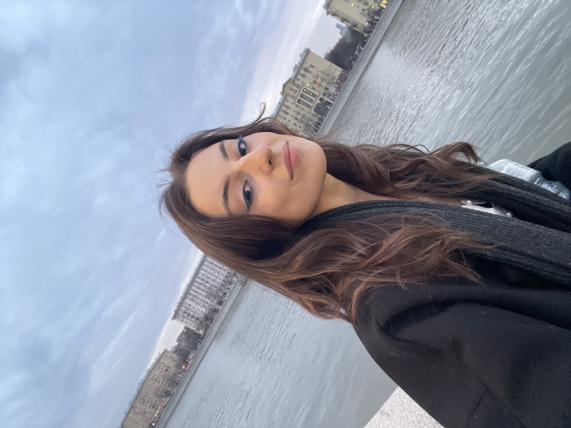 A selfie shows a woman with brown hair, a black jacket, and blue mascara on her eyelashes. She stands along a river in Moscow, Russia. Multi-story buildings line the riverfront in the background. The sky is overcast.