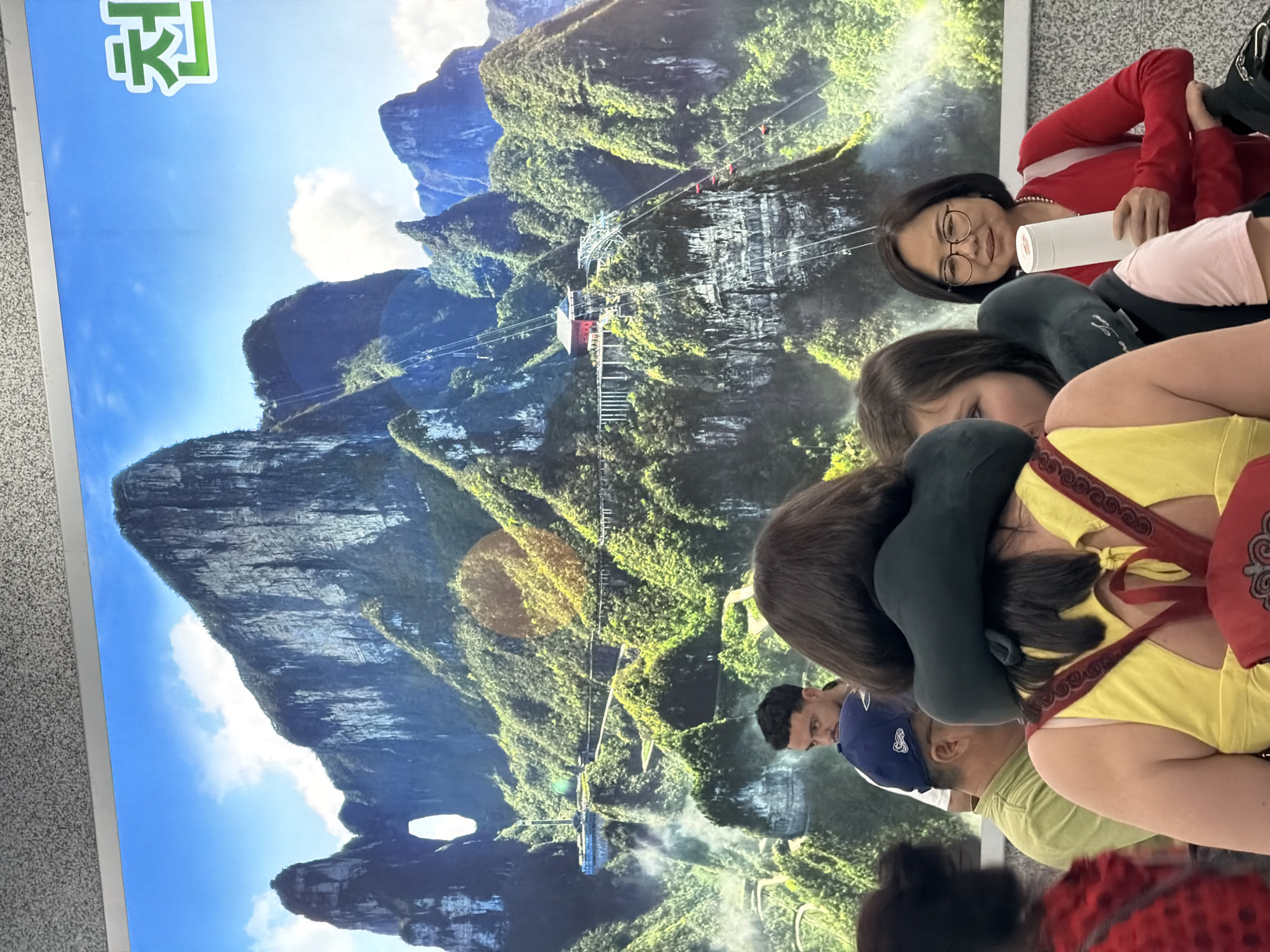 The image depicts a group of people in front of a large poster displaying a mountainous landscape with a cable car system. This landscape is likely Tianmen Mountain, a prominent landmark in Zhangjiajie, China, known for its unique rock formations, the "Heaven's Gate" cave, and a lengthy cable car ride.
Several people are situated in the foreground, partially obscuring the poster. A woman wearing glasses is looking towards the camera, holding a white tube. Another person is wearing a neck pillow. Other individuals are partially visible, their bodies and clothing suggesting a mix of casual attire and possible souvenir purchases. The overall impression is that the people are either waiting or resting near the display. The poster serves as a backdrop, showcasing the scenic beauty of Tianmen Mountain, including the cable car system that connects the base to the summit.