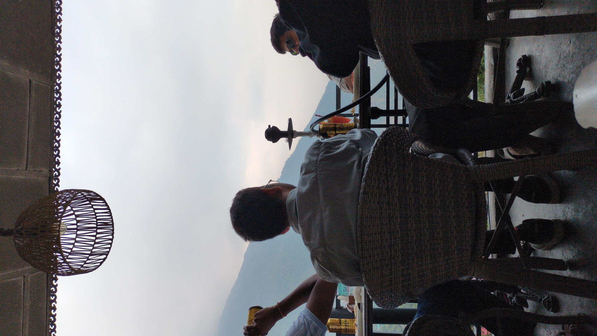 The image depicts a scene in Dharamkot, India. Two individuals are visible; one is sitting and holding a can, while the other is standing. A hookah is situated between them, suggesting shared activity. A rattan chair is prominently featured, and a wicker basket-style lamp hangs from the ceiling. In the background, mountains are visible under an overcast sky.