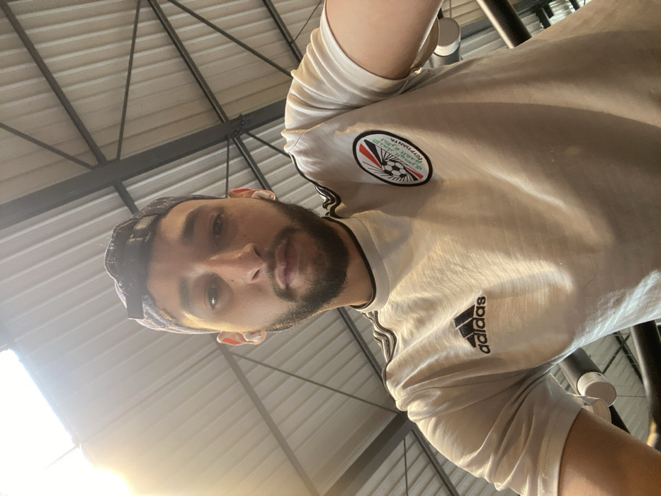 One male individual is depicted in a close-up, upward-angled self-portrait. He has short dark hair, a beard, and light-toned skin, and is looking directly into the camera. He is wearing a white short-sleeved t-shirt with a black Adidas logo on the right chest and a circular emblem on the left chest. The emblem contains a stylized soccer ball, text, and colors resembling the Egyptian flag, with the word "EGYPTIANYA" partially visible. Black stripes are present on the shoulders of the shirt. He is also wearing a dark-colored baseball cap reversed on his head and white wireless earbuds in both ears. The background consists of an indoor setting featuring a corrugated white ceiling with visible dark metal support beams and trusses. A bright light source is present in the upper left portion of the frame. The specific location within the city of Hœnheim, France, cannot be determined from the visual information provided.