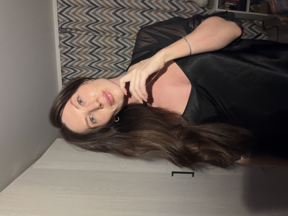 A woman with long brown hair and fair skin lies on a white surface in a black silk robe with lace detailing. She has blue eyes, a full face, and is wearing a thin silver bracelet. Her head is tilted, and she is looking directly at the camera with her hand near her face. The background features a neutral zig-zag pattern. The lighting is soft, emphasizing the smoothness of her skin and robe.
