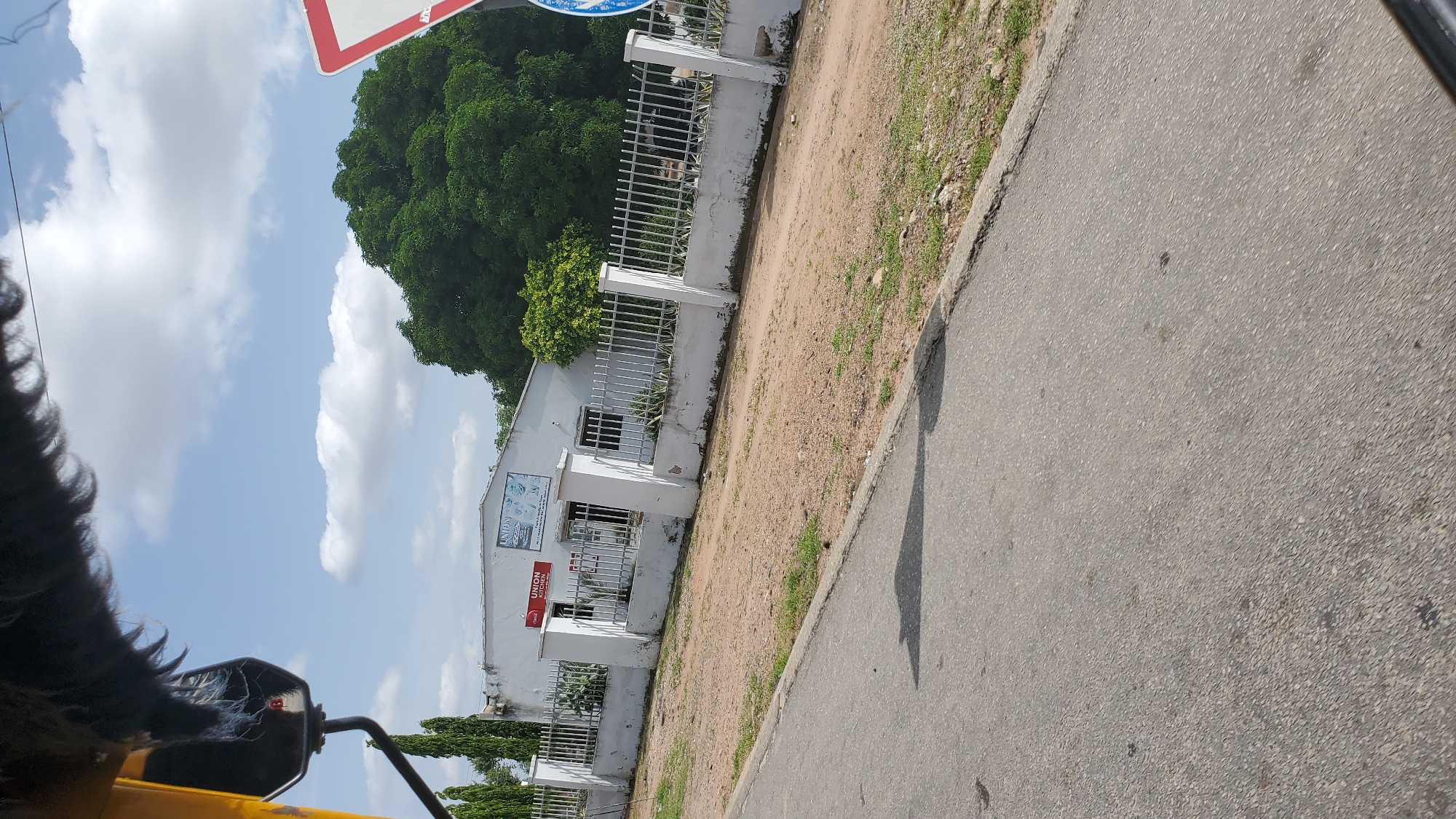The image shows a street view in Jalingo, Nigeria, featuring a white building with a fence in front of it. The building has two signs, one red and one blue, with "UNION KITCHEN" and "UNION" written on them respectively. A large green tree is visible behind the building, and a portion of a yellow vehicle, likely a trishaw or motorcycle with a sidecar, is in the foreground, obscuring the top left corner of the image. A traffic sign, a red and white triangle, is partially visible on the right side of the image. The ground is covered with dirt and grass, and a paved road runs in front of the building. The sky is blue with scattered clouds.