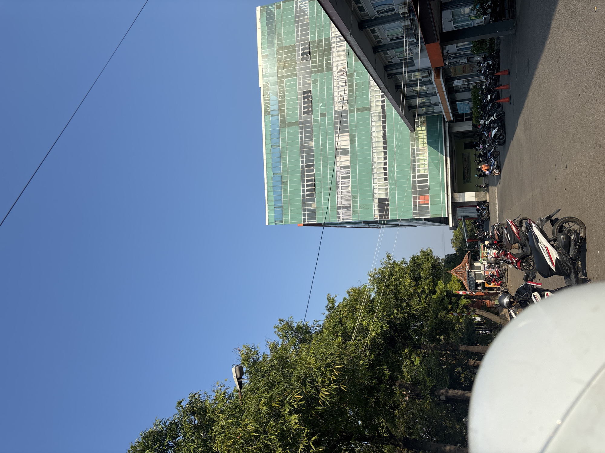 The image captures a sunny day in Cirebon, Indonesia, likely in a public or institutional area, evidenced by a large, multi-story building with a green glass facade and an adjacent, lower building with outdoor areas. A significant number of parked motorcycles are visible in front of these buildings, suggesting a place with high foot traffic or a parking facility. Trees line the left side of the frame, casting shadows on the paved ground, and a streetlamp is mounted on one of the trees. In the foreground, the edge of a white motorcycle helmet is prominent. The sky is clear and blue.
