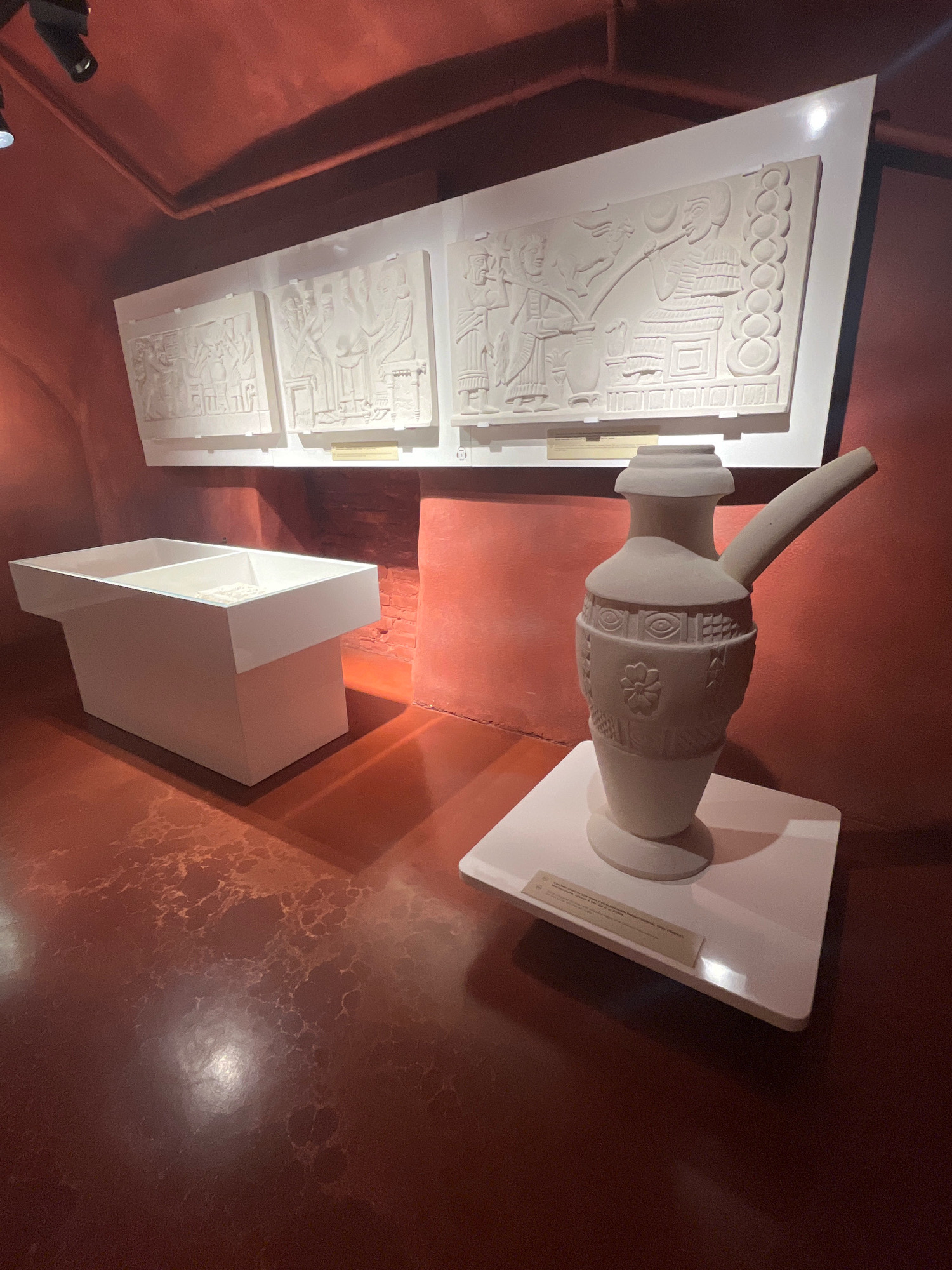 The image depicts an exhibition display within a museum. In the foreground, a pale beige, ceramic pitcher with a long, narrow spout and decorative embossed patterns, including what appears to be a flower and eye motifs, rests on a white rectangular pedestal. The pedestal has a small informational plaque attached to its front edge. Behind the pitcher, a white, rectangular display case with a bifurcated top is positioned. The case contains what appears to be archaeological fragments.
In the background, three rectangular white panels are mounted on a wall, each featuring bas-relief carvings in a pale color. These carvings appear to depict scenes with figures and architectural elements, suggesting historical or mythological narratives. The walls of the exhibition space are a deep reddish-orange hue. The floor is a similar reddish-brown color, with a textured, somewhat mottled appearance. Two black spotlights are visible in the upper left corner of the image, illuminating the display. The scene is lit with dramatic lighting, creating shadows.
There are no people visible in the image. The overall impression is of an archaeological or historical exhibit. The specific location in Lviv, Ukraine, is not evident from the visual information provided in the image.