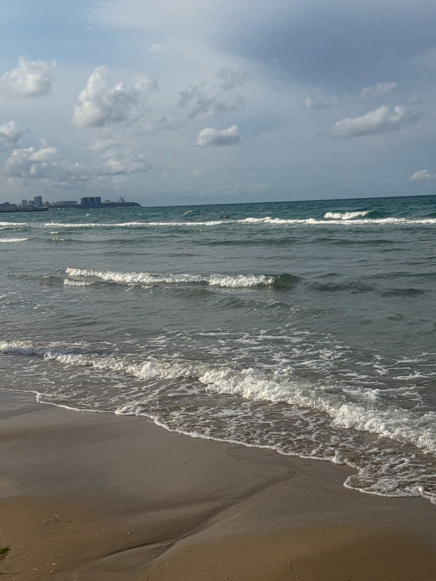 The image captures a scene at a beach in Anapa, Russia, with the Black Sea in the foreground and a cityscape visible on the horizon. The sky is overcast with scattered clouds, casting a muted light on the scene. Gentle waves are lapping at a sandy shore, creating white foam as they recede. In the distance, buildings and structures of a city line the coast. There are a few small figures, presumably people, visible in the water, suggesting recreational activity. The overall impression is a calm, coastal environment with a settled weather pattern.