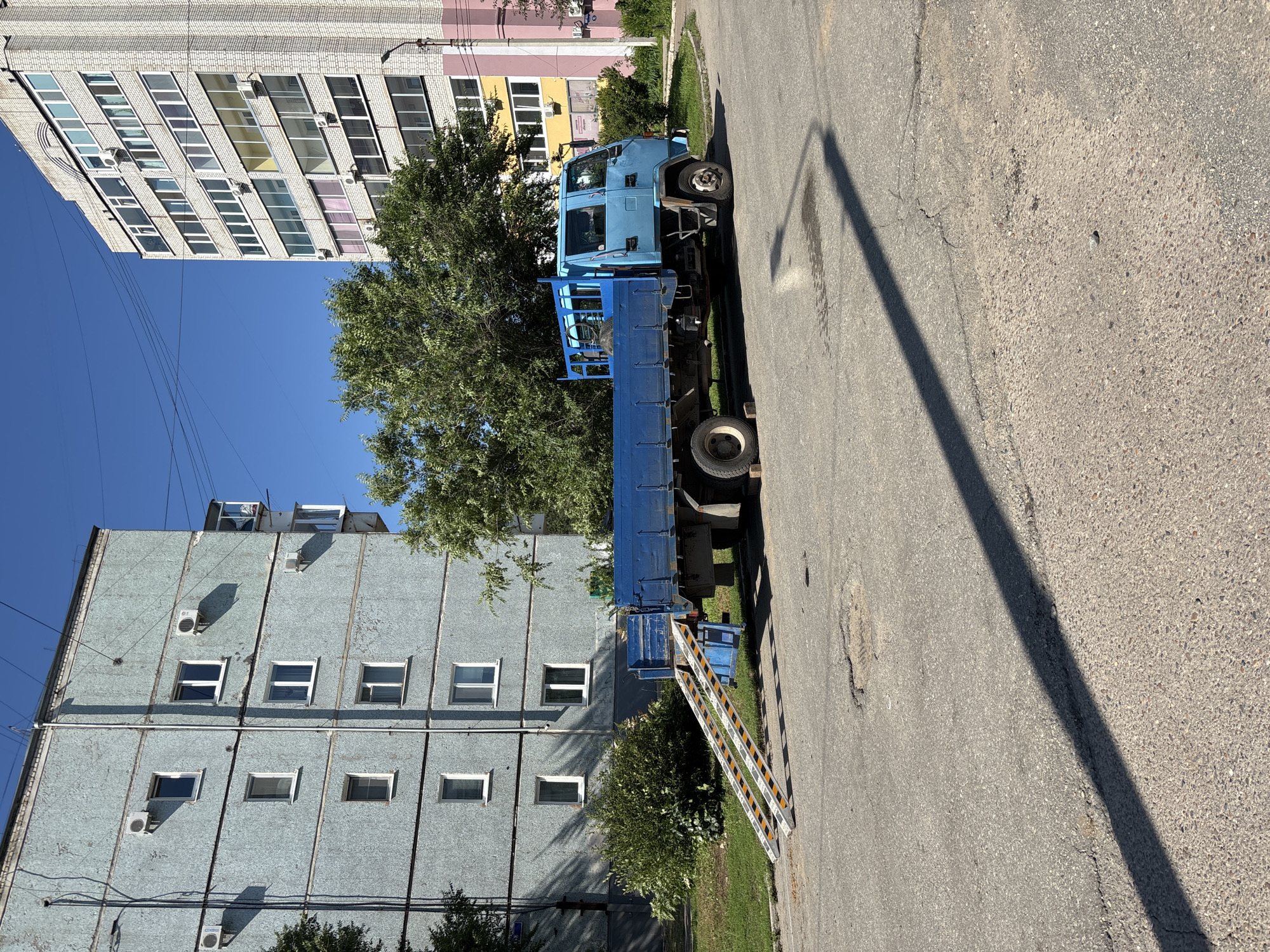 A bright blue flatbed truck with two ramps extended from its bed is parked on an asphalt surface in front of two apartment buildings. The truck is positioned at an angle to the buildings, with the ramps leading towards the ground. The building on the left is a multi-story structure with a textured, pale blue facade, featuring several windows and air conditioning units. The building on the right is taller and clad in white brick, with numerous windows, balconies, and colorful accents on its lower levels. Trees and bushes are visible between the truck and the buildings, and the sky is clear and blue. The ground shows signs of wear and tear, with some cracks and potholes in the asphalt, and a prominent shadow of a lamppost falls across the scene. The scene appears to be in a residential urban area.