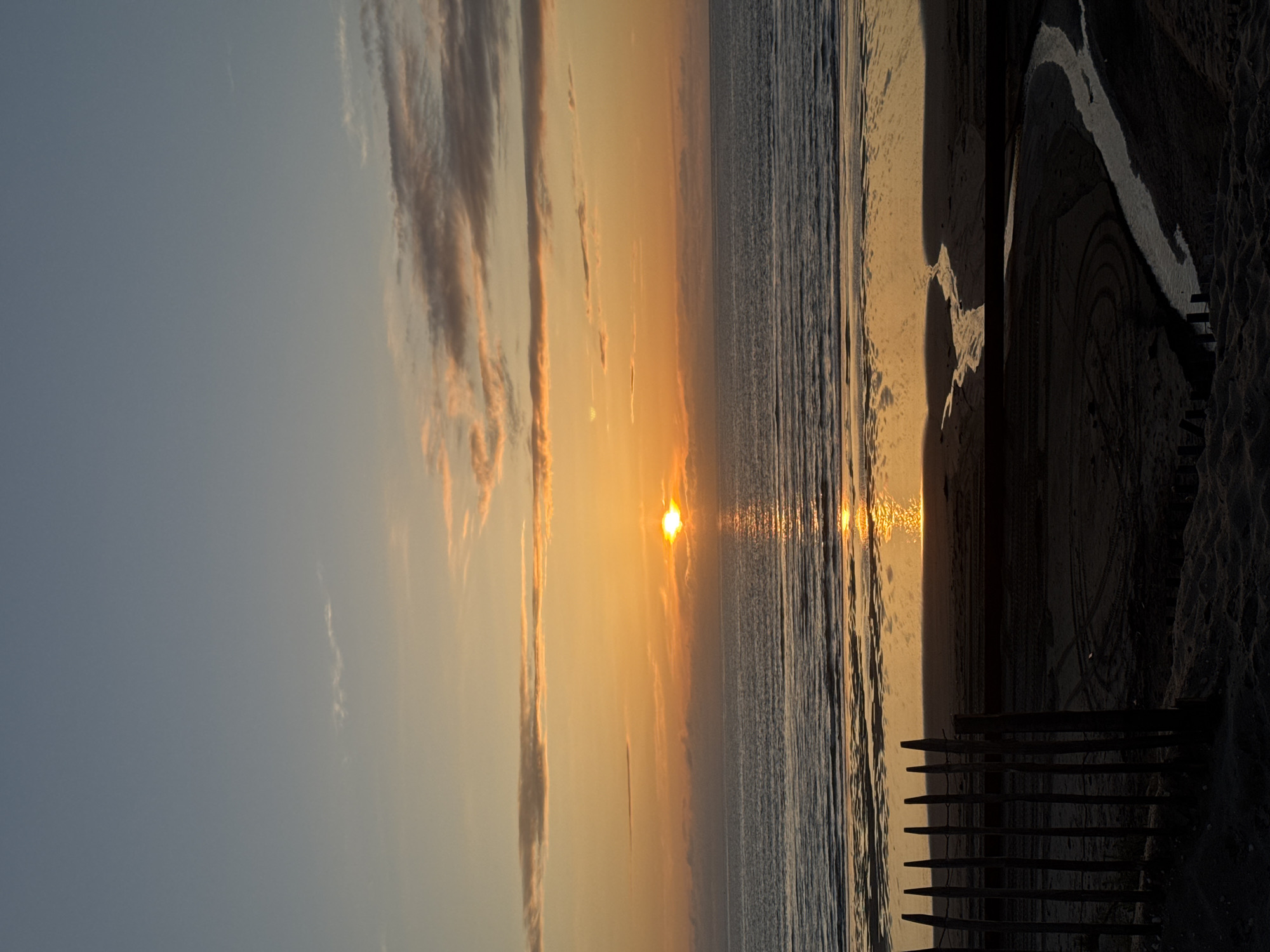 The image depicts a sunset over a beach. The sun is setting on the horizon, casting an orange glow on the water and clouds. There are long clouds in the sky. The beach is sandy, and there is a wooden fence in the foreground. There is also a stream cutting through the sand. This is likely a beach in Soulac-sur-Mer, France.
