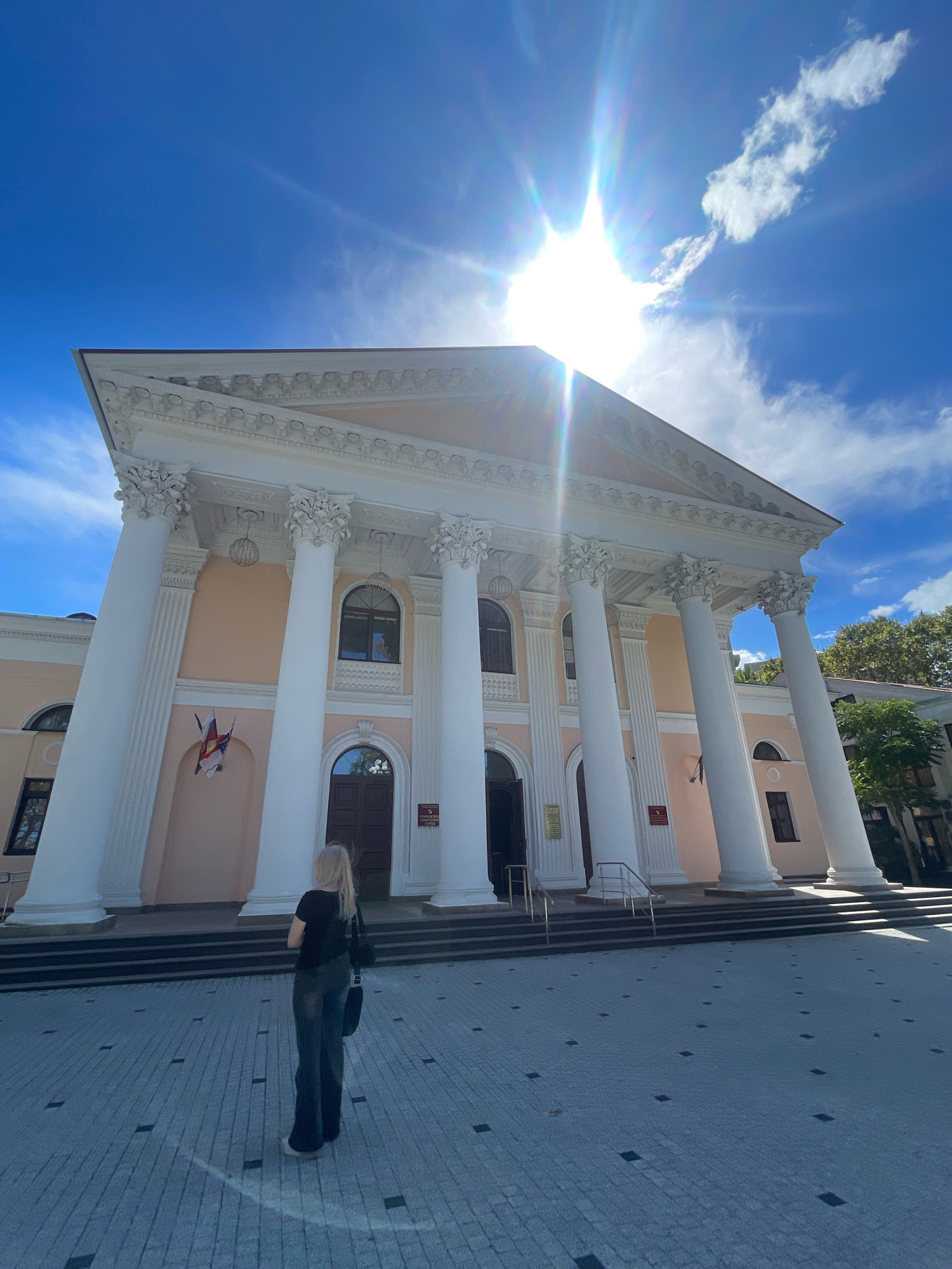 The image captures a low-angle, wide shot of the Winter Theatre in Sochi, Russia, on a sunny day with a bright blue sky and scattered clouds. The sun is prominently positioned at the top of the frame, causing lens flare and washing out parts of the sky and the building's facade. The theatre itself is a grand neoclassical structure with a pale peach exterior, adorned with numerous tall, fluted white columns supporting an ornate entablature. There are several arched windows and doorways visible on the building's front. Two flags are displayed near the central entrance. In the foreground, a young woman with blonde hair, wearing a black top and dark, wide-leg trousers, stands with her back to the camera, facing the theatre. She is positioned on a paved plaza made of grey interlocking bricks with square dark inserts, creating a patterned surface. The plaza slopes slightly away from the building. The overall impression is one of architectural grandeur under a bright, clear sky.
