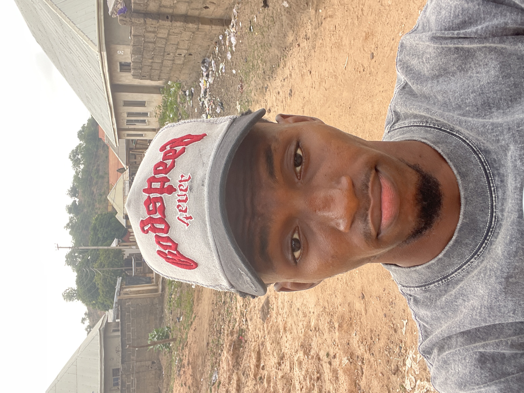A young Black man is featured in a close-up, outdoor portrait, looking directly at the camera with a neutral expression. He is wearing a light grey baseball cap with red embroidered text on the front that reads "Godspeed fever". His attire also includes a grey, possibly acid-washed or textured, t-shirt. He has a short beard or stubble on his chin.
The background reveals an unpaved, sandy-brown ground with scattered litter and debris. Beyond this, several modest buildings are visible under an overcast sky, suggesting daytime conditions. These structures feature corrugated metal roofs and walls of various materials, including what appears to be concrete blocks. One building in the mid-ground has a faint "02" or "OZ" marked on its facade. Green trees and distant hills are visible behind the buildings, indicating a vegetated landscape. The setting appears to be a residential or semi-urban area, consistent with a location like Lokoja, Nigeria.