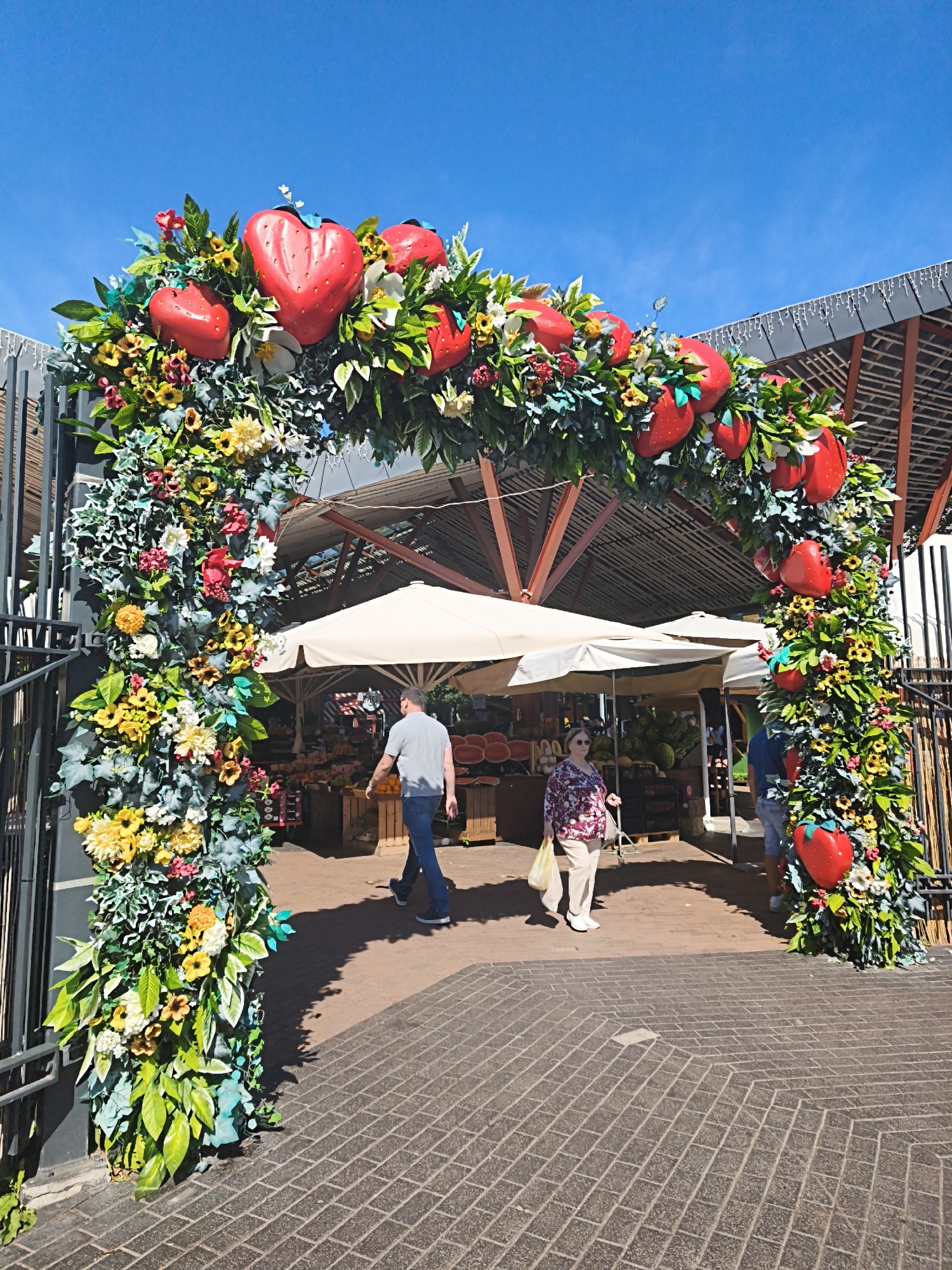 This is a daytime, eye-level, full shot image of a market entrance decorated with a floral arch. The arch is adorned with large, red, strawberry-shaped decorations and various artificial flowers and greenery, including yellow and white flowers. The arch is set against a clear blue sky. In the background, a market structure with a dark roof and brown support beams is visible. Several white market umbrellas are set up, and stalls with fruits and vegetables are partially visible.
In the foreground, two people are walking under the arch. On the left, a man wearing a grey t-shirt and blue jeans is walking away from the camera. On the right, a woman wearing a floral blouse and light-colored pants is walking towards the camera, carrying a plastic bag. The ground in front of the arch is paved with brown bricks laid in a herringbone pattern. The overall scene suggests a vibrant outdoor market environment. The location is not specified in the image, but it is presented as being in Tallinn, Estonia.