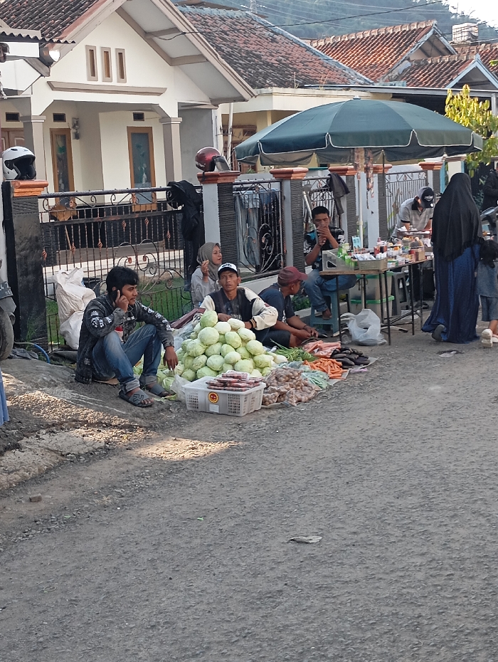 The scene is a street market in the city of Kutawaringin, Indonesia, located at the coordinates -6.974, 107.519. There are several vendors selling produce and other goods from stalls on the side of the road. A man is sitting on the ground next to a pile of cabbage heads. A few other vendors are sitting or standing in front of their stalls. There are several customers browsing the stalls, and some people are walking by. There are several vehicles parked on the side of the street, including a motorcycle. The environment is a typical urban street scene in a developing country. The day is bright and sunny.