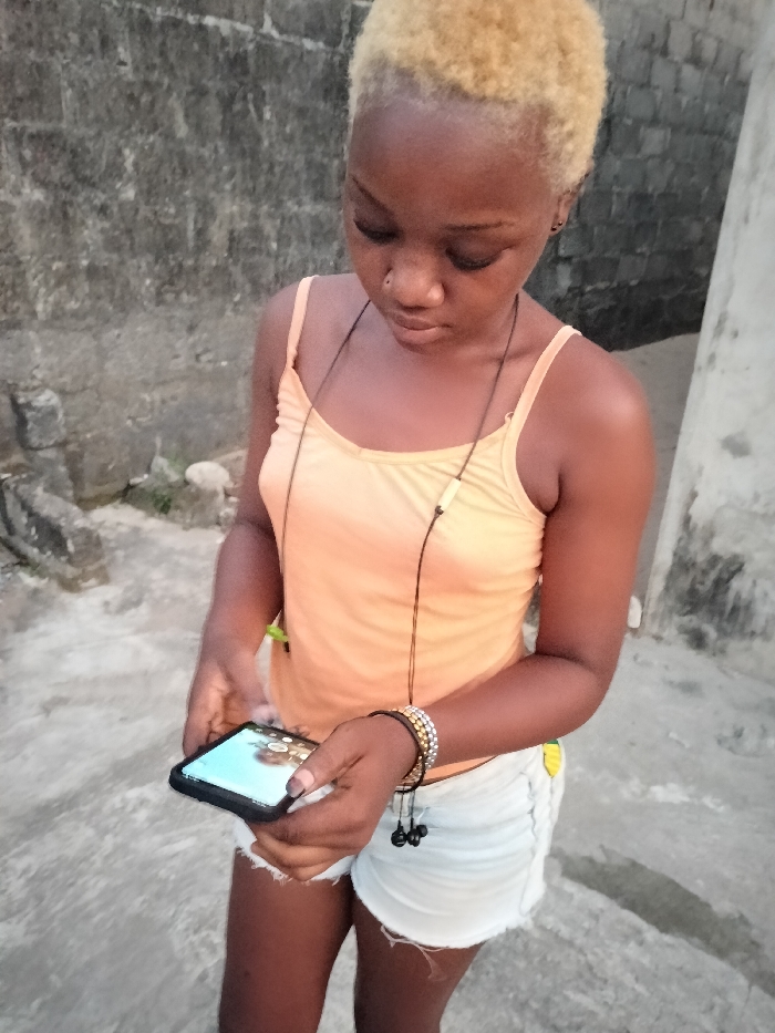 A young woman with bleached blonde hair stands outside, looking down at a phone she holds in her hands. She wears an orange tank top and denim shorts. A necklace hangs around her neck and beaded bracelets adorn her wrist, with earbuds hanging from them. The background shows a concrete wall and paved ground, suggesting an urban setting.