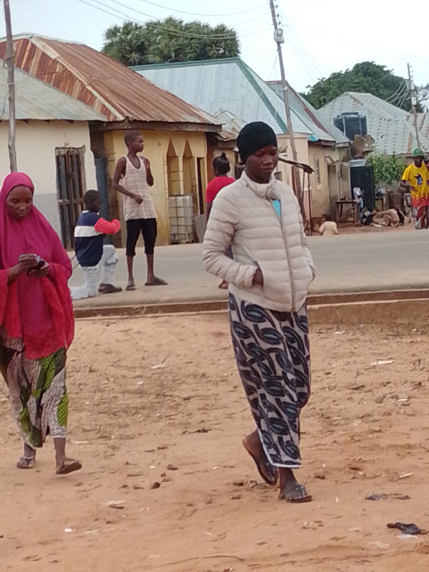 The image depicts a street scene in Tempure, Nigeria. In the foreground, a woman wearing a pink hijab and a patterned skirt is looking at her phone. To her right, a person in a light-colored puffer jacket and a patterned skirt is walking towards the right side of the frame. Behind them, a young boy in a blue and white shirt and white pants is kneeling. Further in the background, other individuals and various buildings with corrugated metal roofs are visible. The ground is a dusty, reddish-brown color, indicative of a dry environment. Power lines are strung across the sky, suggesting an inhabited area. The overall atmosphere appears to be that of a typical day in a Nigerian town.