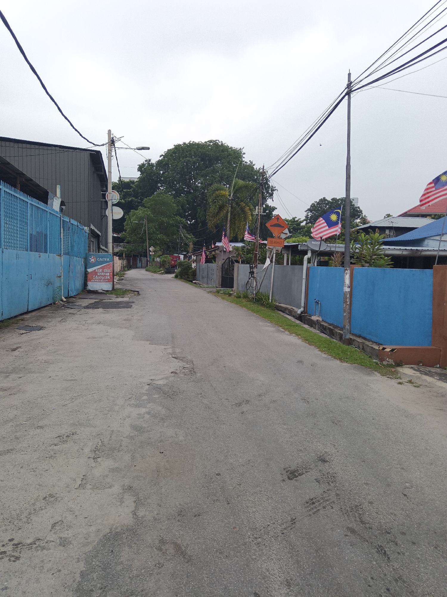 The image depicts a street scene in Kuala Lumpur, Malaysia. On the left, a large blue metal fence runs along the side of a building with a gray corrugated metal exterior. A Caltex sign advertising "Pusat Servis & Minyak Berkualiti" and "Tyre Service" is propped up against the fence. The street itself is paved with asphalt and shows signs of wear and tear, with tire marks visible in places. Several Malaysian flags are displayed on poles along the right side of the street, fluttering in the breeze. A speed bump sign with "35 Km/h" is also visible. Electrical wires crisscross overhead, supported by utility poles. The sky is overcast, suggesting a cloudy day. Houses with blue roofs and gray walls are visible in the background, interspersed with trees and vegetation. The scene appears to be a residential or mixed-use area in a developing urban environment.