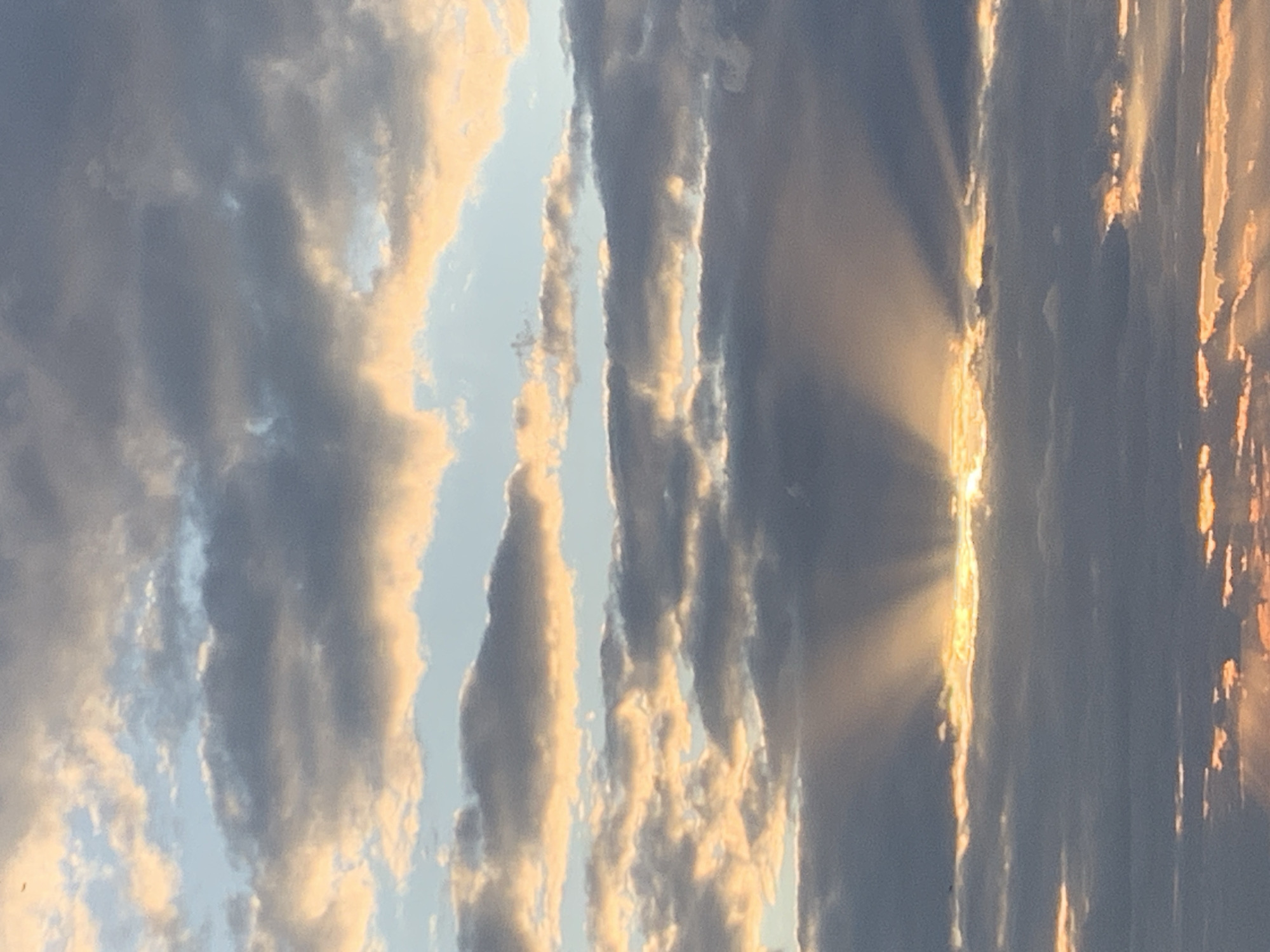 The image shows a cloudy sky with crepuscular rays emanating from behind the clouds. The sun appears to be low on the horizon, casting an orange and yellow glow on the clouds. There are no people or objects visible in the image. The location is not specified in the image.