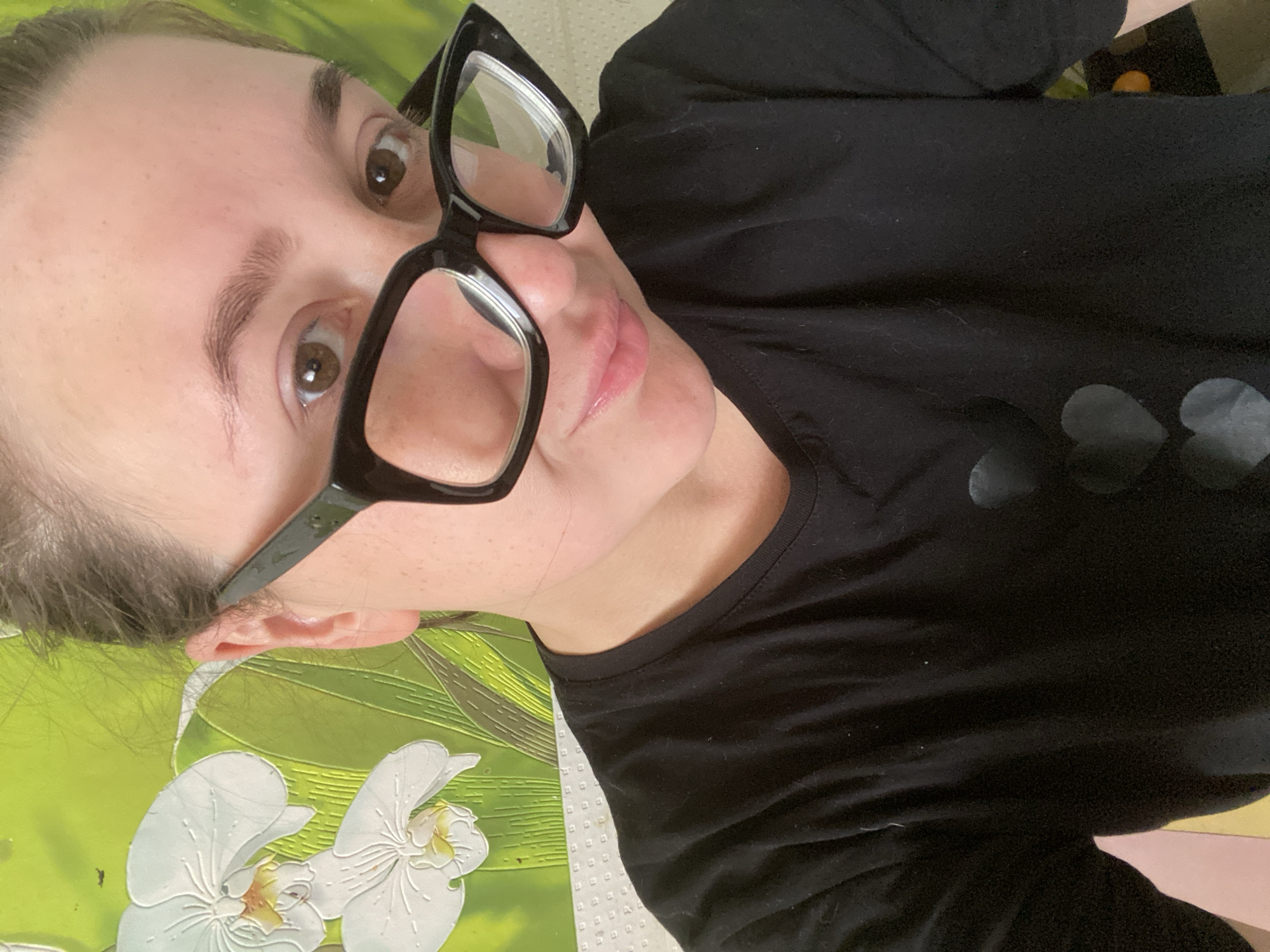 A close-up, selfie-style photograph shows a person wearing black-rimmed glasses and a black t-shirt with heart designs. The person has brown eyes and light skin with freckles. They are looking at the camera with a slight smile. The background is a green wall with a floral pattern, possibly depicting orchids. The lighting suggests it is daytime. The location in Chaikovsky, Russia, is not identifiable from the image alone.