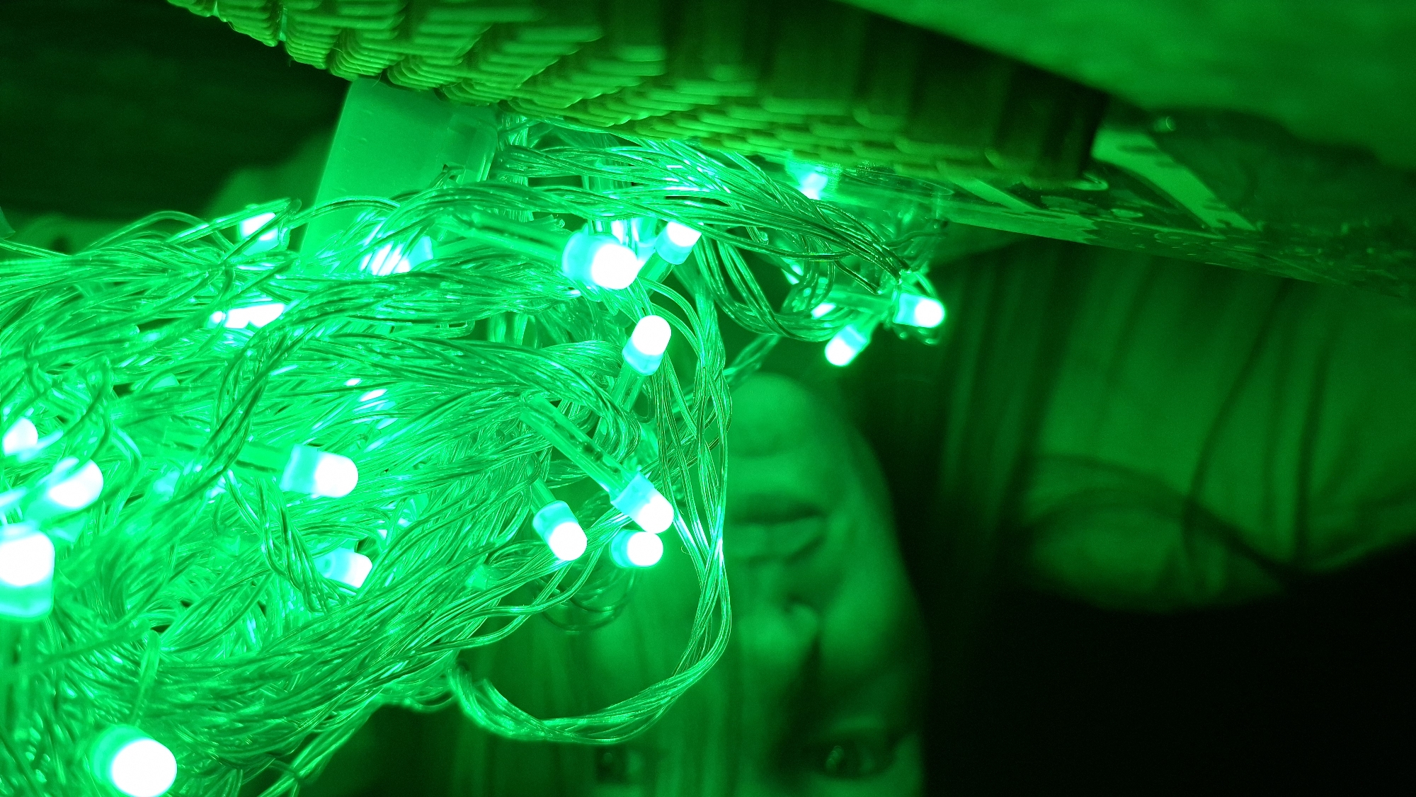 The image displays a close-up view of numerous green LED string lights, illuminated brightly against a dark background. The lights are intertwined, forming a dense tangle of thin green wires and glowing bulbs. Each bulb emits a vibrant green light, making the wires appear translucent and enhancing the overall green hue of the scene.
In the upper portion of the image, a woven texture, possibly from a basket or a piece of furniture, is visible, partially lit by the green glow. Beneath and behind the cluster of lights, a human face is faintly discernible. It is highly out of focus and bathed in the intense green light, with only hints of lips and possibly an eye or brow evident. The presence of the face suggests a person is either interacting with or situated very close to the lights.
The setting appears to be dark, likely nighttime or a dimly lit indoor space, as the green lights are the primary source of illumination. There is no specific activity depicted beyond the lights being turned on, creating an atmospheric and somewhat mysterious ambiance. No text is visible in the media file. The location context of Большой Букор, Russia, is provided, but no specific visual cues within the image confirm this geographical detail.
