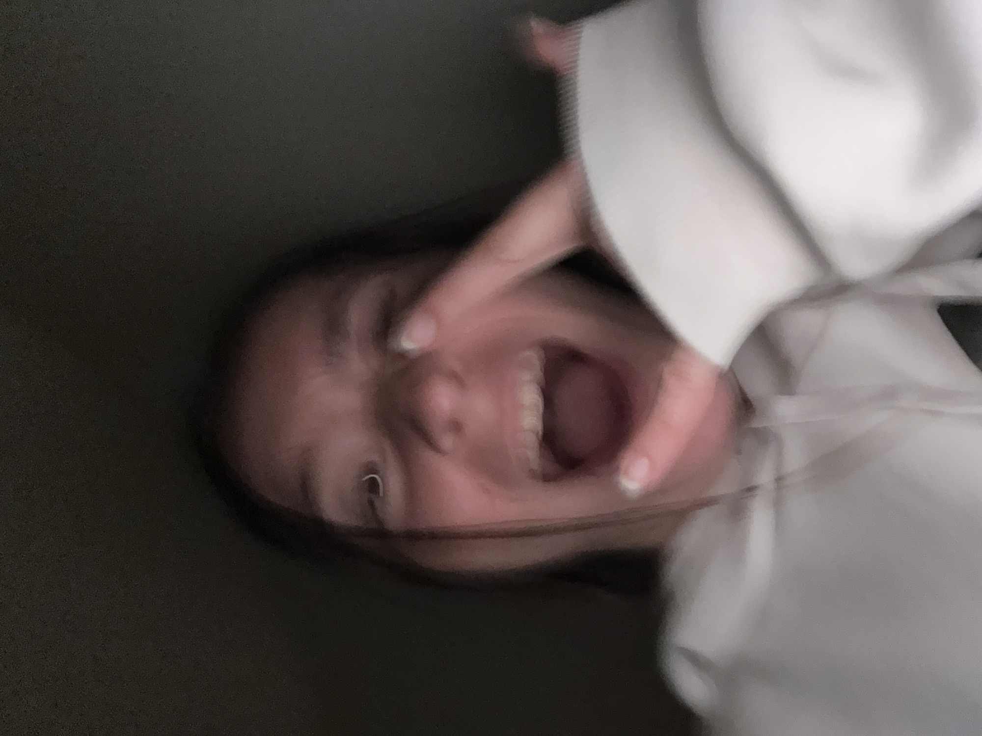 This is a close-up, blurry, portrait-style image of a young person. The person is making a peace sign with their right hand, with their index and middle fingers extended. Their mouth is open in a wide smile or laugh, and their teeth are visible. They are wearing a light-colored hooded sweatshirt. The background is dark and indistinct, suggesting an indoor setting with low lighting. There is no discernible location in Sofia, Bulgaria.