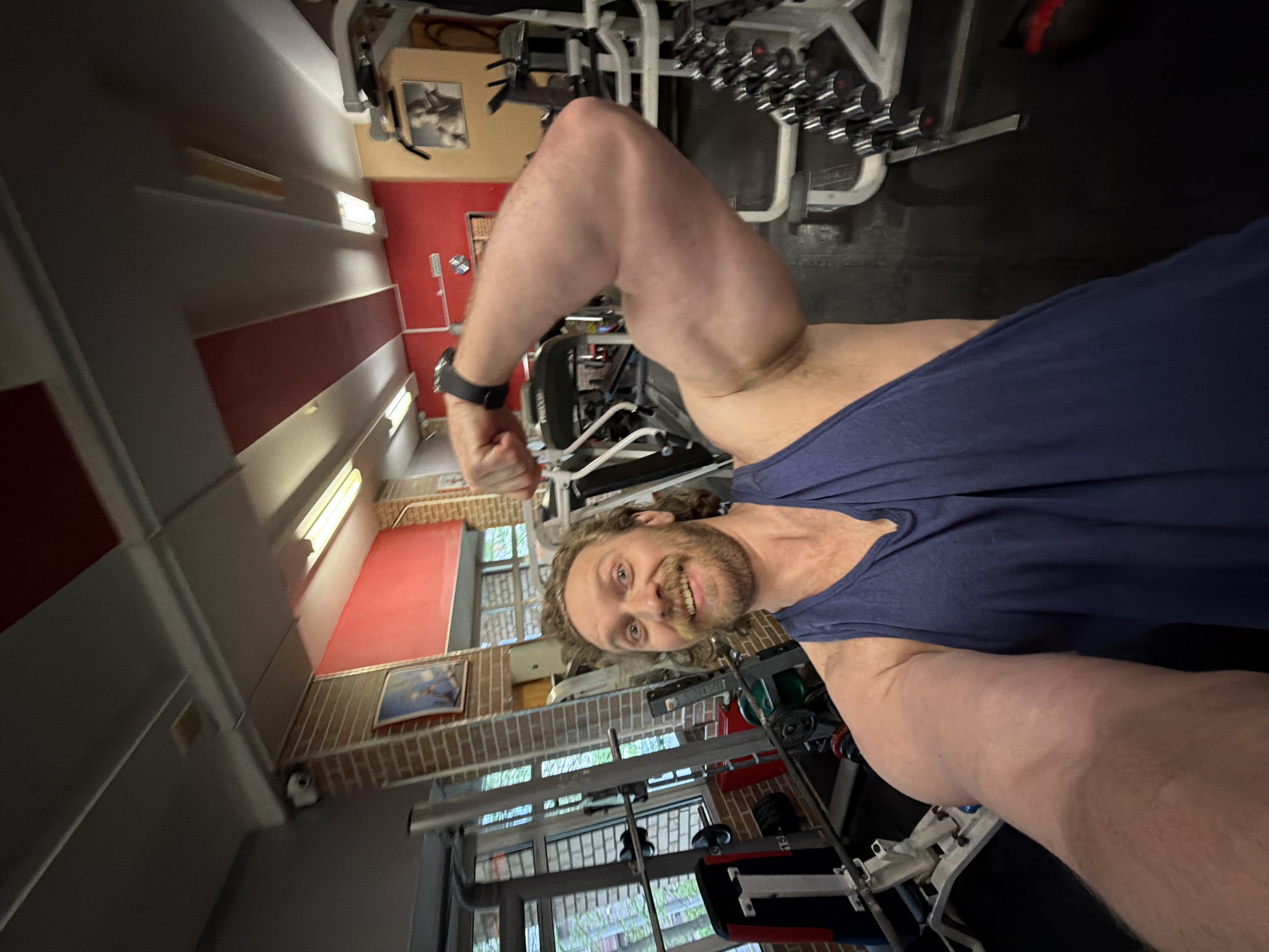 A man is posing in a gym while flexing his bicep. The gym features weightlifting machines and dumbbells. Walls are visible, with some painted red and others with a brick pattern. The location cannot be determined to be in Saint Petersburg, Russia based on the visual information provided.