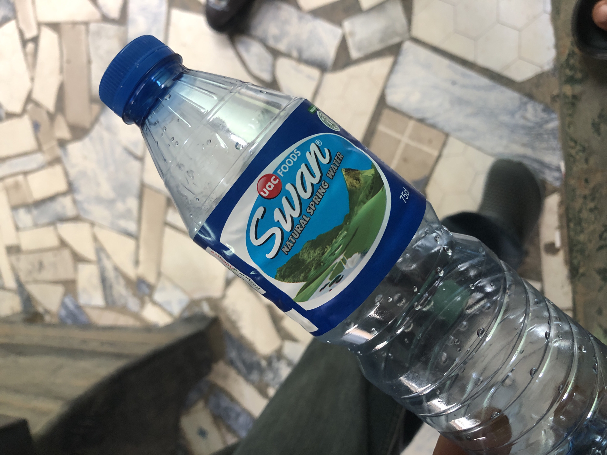 A close-up, angled shot shows a hand holding a clear plastic bottle of Swan Natural Spring Water. The bottle is partially filled with water and condensation is visible on its surface. The blue label features a landscape image with green hills and a body of water, and the brand name "Swan" in white, stylized font, along with "UAC FOODS" in smaller red text. The text "NATURAL SPRING WATER" is also visible. The bottle cap is blue. The background appears to be an outdoor tiled surface with a mosaic pattern of light and dark grey, beige, and white tiles. Part of dark green fabric, possibly clothing, is visible on the left side of the frame. The lighting suggests it is daytime.