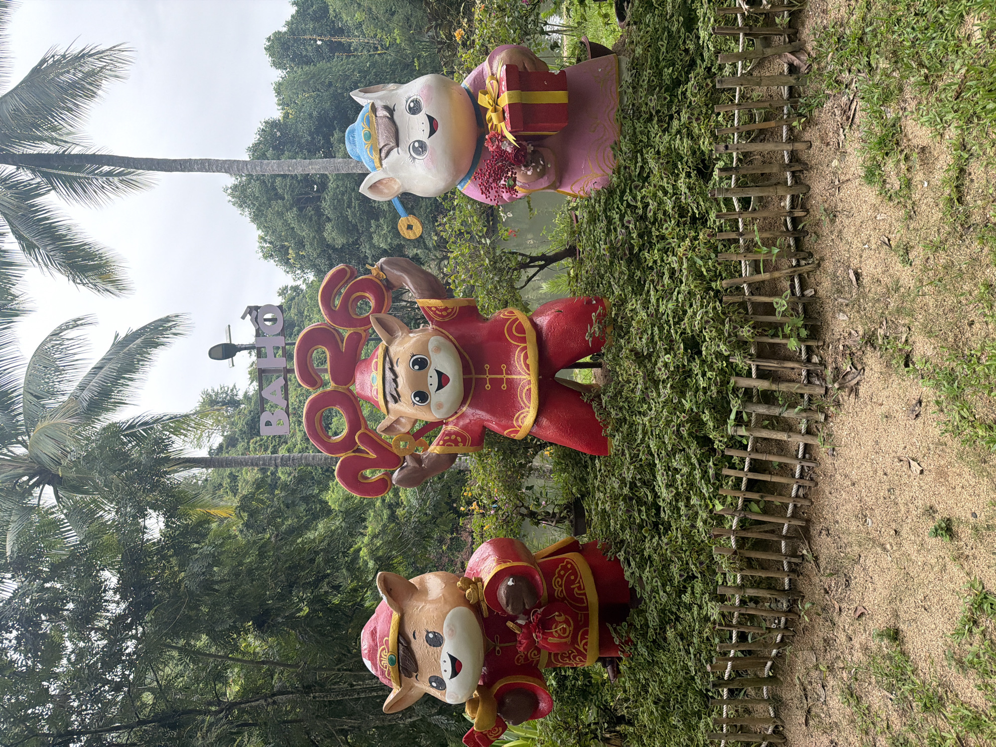 In Nam Ninh Hoa, Vietnam, a trio of cartoonish ox statues are displayed outdoors. The statues are dressed in traditional red and gold attire, symbolizing good fortune and prosperity. The central ox proudly holds aloft the numbers "2026" in large red digits. To its left, another ox clutches a bundle of gold coins and a red pouch, while the ox on the right holds a bouquet of red flowers and a wrapped gift. They stand in front of lush green foliage, with palm trees and a body of water visible in the background. A small, rustic bamboo fence separates the sandy ground in the foreground from the landscaped area where the statues are placed. The scene appears to be a festive decoration, possibly for a holiday celebration.