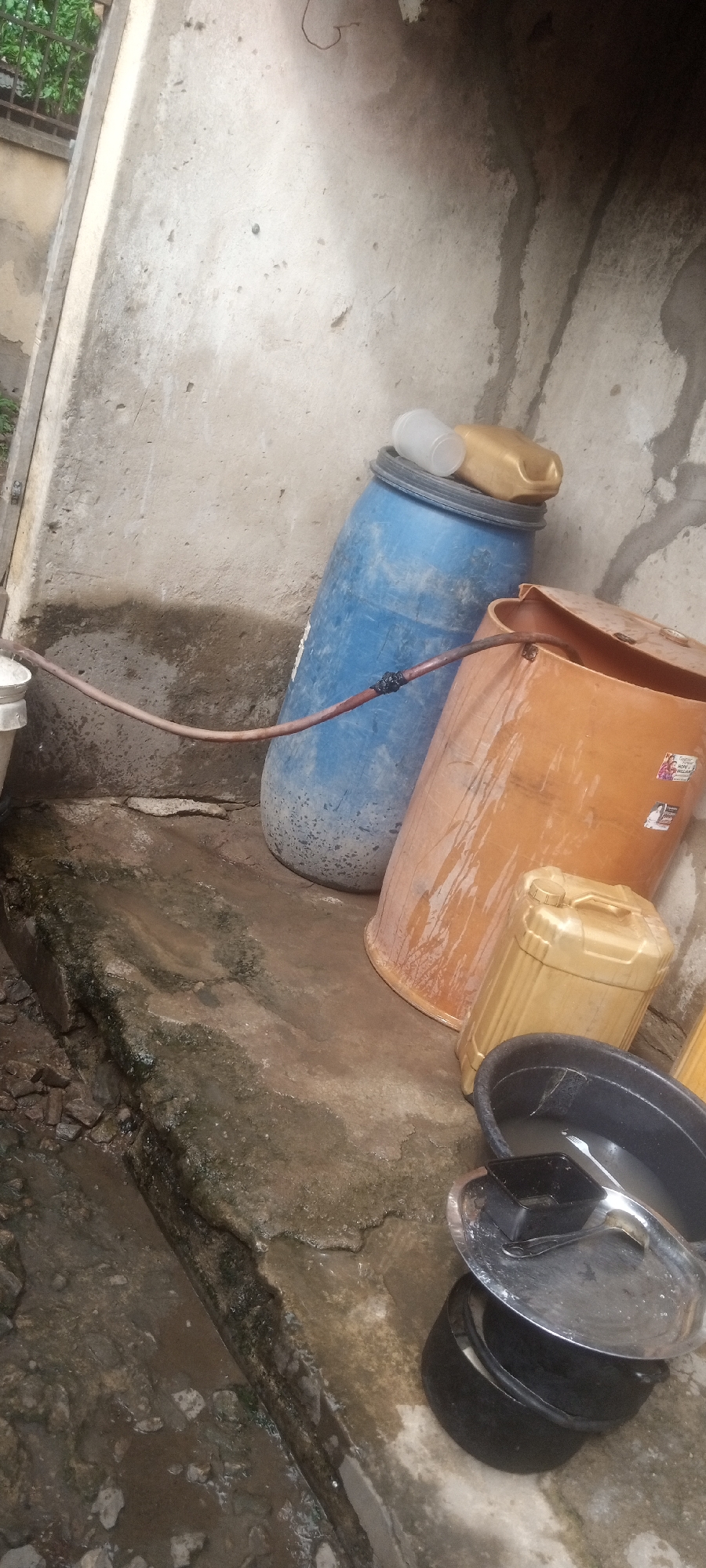 The image depicts an outdoor scene in Sabon Tasha, Nigeria. A blue plastic barrel and an orange plastic barrel are leaning against a concrete wall. A brown hose connects the blue barrel to a white pipe on the left. The ground is wet and appears to be concrete. An orange plastic jerrycan is next to the orange barrel. A black basin and a pot with a lid and utensils are in the foreground. The setting appears utilitarian, possibly a backyard or service area.