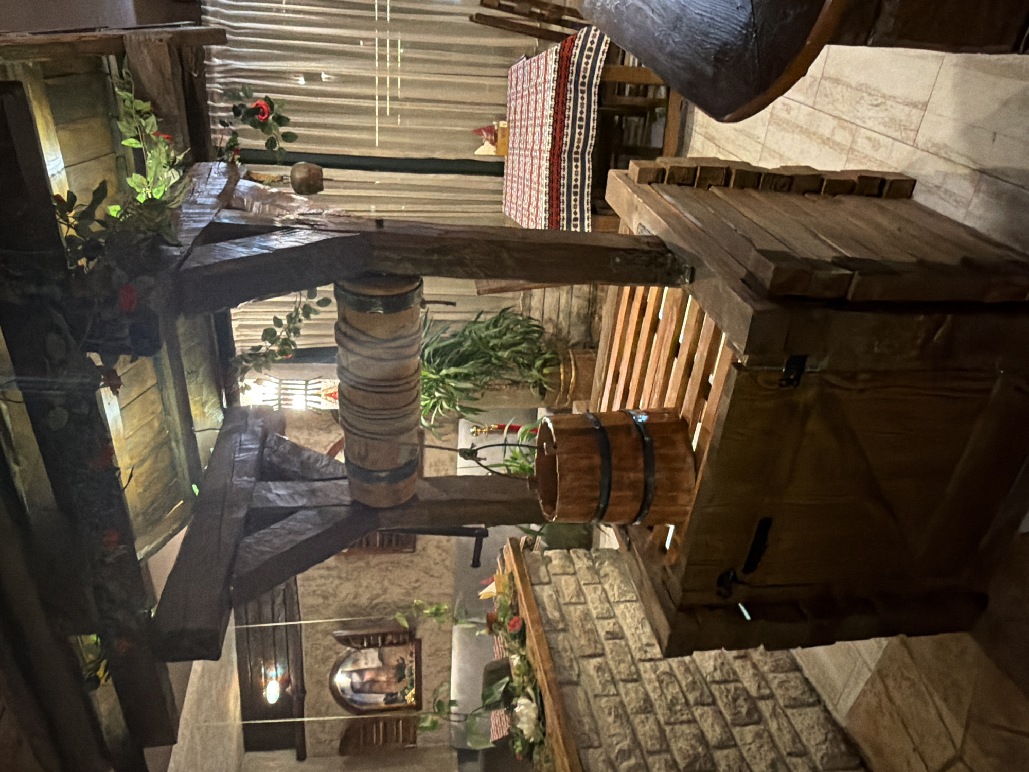 The image shows an indoor decorative well structure made of wood in a dimly lit restaurant or cafe. The well has a wooden bucket suspended from a rope and a crank mechanism. The structure is detailed with wooden planks and beams, and decorative vines with artificial red flowers are draped around it. In the background, there is a table set with a red and white patterned tablecloth, chairs, and a curtained window. To the left, a stone counter with plants and a painting on the wall is visible. The overall ambiance is rustic and possibly themed. This scene is located at a restaurant in Kumbor, Montenegro.