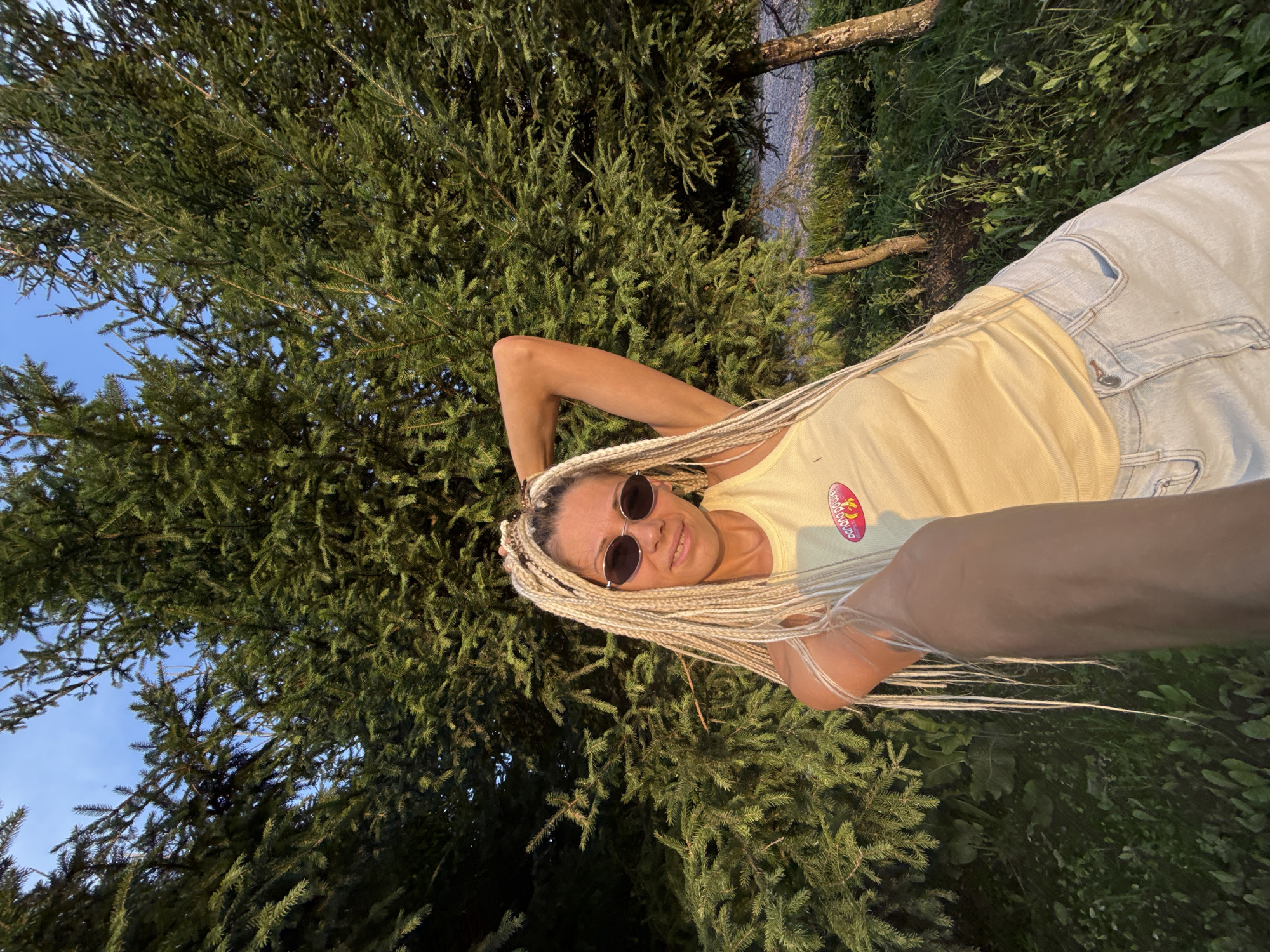 In Zhukovo, Russia, a woman with blonde braids and sunglasses takes a selfie in front of dense evergreen trees. She is wearing a yellow tank top with a graphic print and light-wash jeans. The setting appears to be outdoors, with a clear blue sky visible through the branches and green foliage on the ground. The lighting suggests it is either early morning or late afternoon.