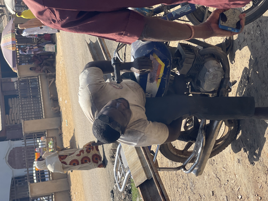 The image depicts a street scene in Zaria, Nigeria, under bright daylight. In the immediate foreground, a dark-skinned male, wearing a dirty white short-sleeved t-shirt, dark trousers, and sunglasses pushed onto his forehead, is bent over a blue motorcycle. His left hand is positioned on the motorcycle's fuel tank area, which exhibits wear and a partial white, yellow, and blue stripe design. The motorcycle has a visible engine block and exhaust pipe, and a metal rack on its rear carrying a rectangular light-colored object, possibly a piece of wood or metal. To the right, only the torso and arm of another individual, wearing a maroon long-sleeved garment and holding a blue smartphone with a patterned case, are visible. The ground is dusty and unpaved in the foreground.
In the midground, a paved road stretches across the frame. A woman dressed in a cream-colored hijab and a patterned long dress with orange, black, and white motifs walks away from the camera on the road. To the left of the road, a small vendor's cart with various bottled items is visible next to a black metal fence.
The background features a building with a light-colored facade, in front of which large sacks are stacked, likely containing grain or cement. Further behind, under a multi-colored striped umbrella, a group of individuals are seated and standing. Several other people, including women in hijabs and men in traditional attire, are visible walking or standing in the distance, suggesting a busy urban or market environment.