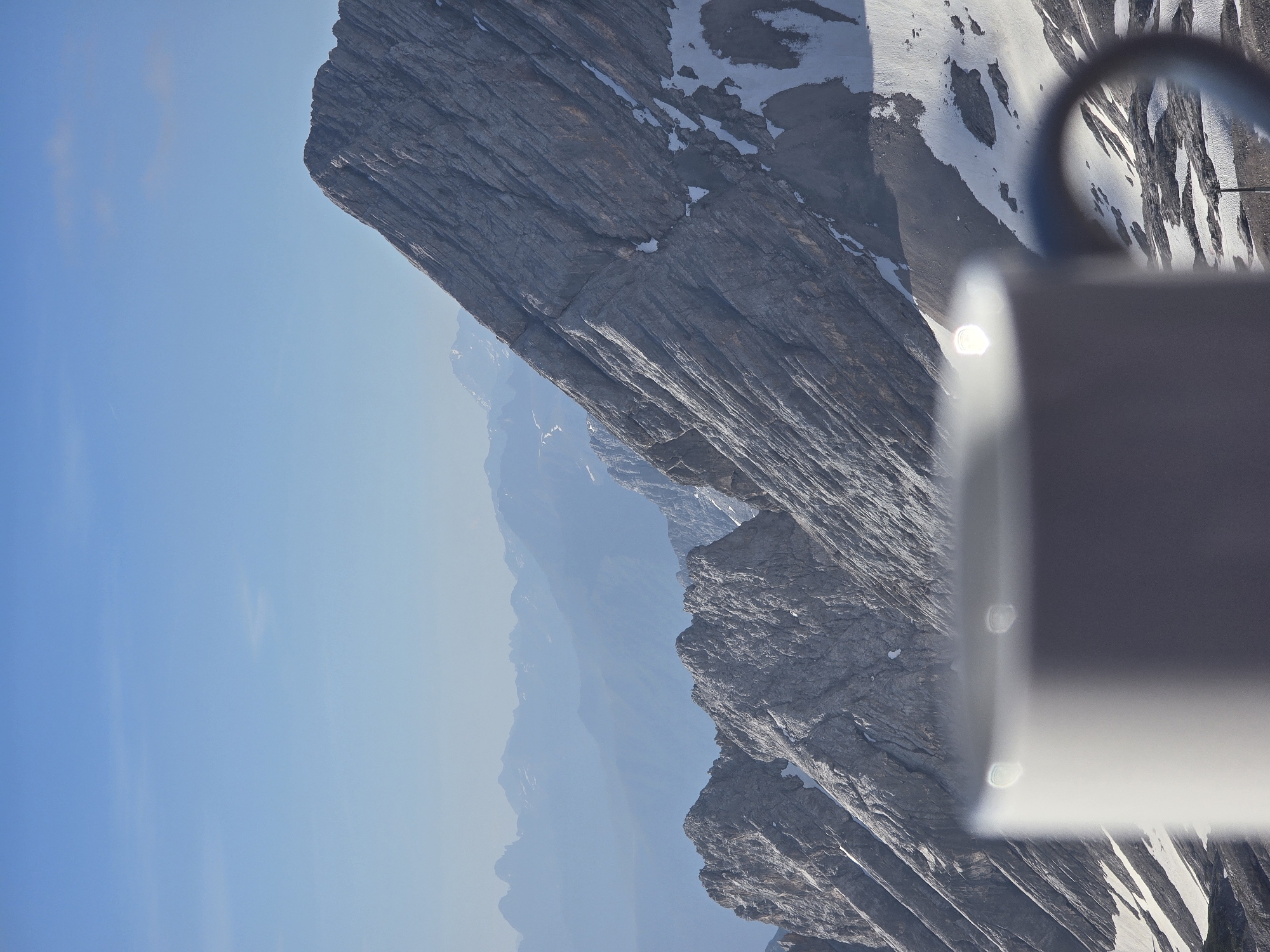 A white mug is in the foreground, out of focus, obscuring the bottom left of the image. The mug has a dark handle visible on the right side. Behind the mug, a vast mountain landscape unfolds under a clear blue sky. The mountains are rugged and rocky, with patches of snow visible on their slopes. In the far distance, several layers of mountains fade into a hazy blue, creating a sense of depth and scale. The scene evokes a feeling of serene elevation, likely captured from a high vantage point such as the Zugspitze, Germany's highest peak, which is located near Garmisch-Partenkirchen.