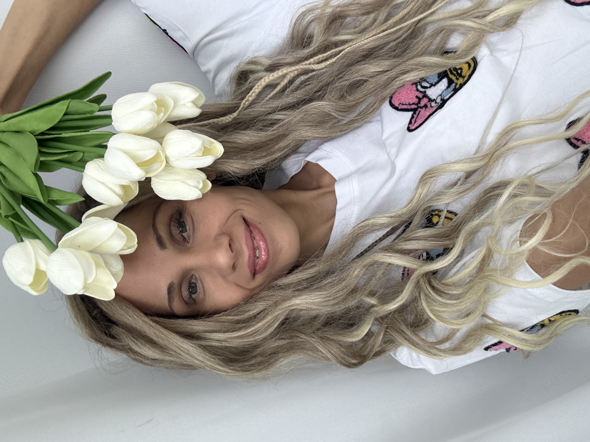 A woman with wavy blonde hair and a braid is lying down, wearing a white t-shirt with butterfly patches. She is smiling and has a bouquet of white tulips near her head. The environment is a white surface. Location information is not discernible from the image.