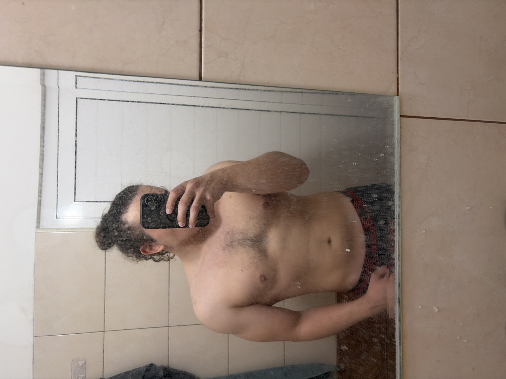 An upright, eye-level mirror selfie shows a person, likely male, with dark, curly hair tied back in a bun. The person is shirtless, revealing a muscular torso with some chest hair and visible abdominal definition, and is wearing patterned dark shorts or underwear with red accents. They are holding a black smartphone with a multi-lens camera setup, which partially obscures their face.
The scene is a bathroom, indicated by the large rectangular mirror and the light brown or beige square tiles with dark grout lines that cover the walls and possibly the floor. A white window frame is reflected in the upper left portion of the mirror. The mirror surface appears slightly smudged or speckled, possibly from water droplets. Faintly visible in the reflection, on the lower left, are a dark, folded towel and clear adhesive hooks on the tiled wall. The activity is the act of taking a mirror selfie. No specific time of day or outdoor weather is discernible. No text is visible in the image. The location context is Plovdiv, Bulgaria.