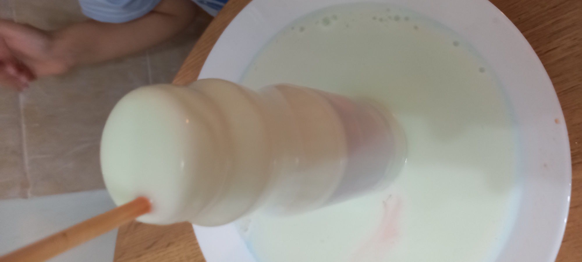 A blurred image captures a dessert being prepared, likely at home, though the exact location in Krasnodar, Russia cannot be determined from the visual alone. A young child's hand and blue-striped sleeve are visible on the left edge of the frame. The central focus is a chimney cake, light-colored and cylindrical with a spiraled texture, suspended in a bowl filled with a milky, off-white liquid. A wooden skewer protrudes from the bottom of the cake. A white bowl containing the liquid rests on a wooden surface. Streaks of pink or red color are visible in the liquid, perhaps from an ingredient used to flavor the cake.