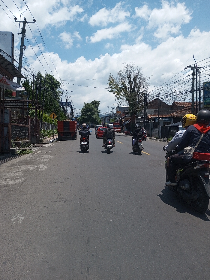 This daytime image captures a street scene in Tagtse Tashidingkha, Bhutan, under a blue sky with scattered white clouds. The asphalt road, which lacks visible lane markings, extends into the distance. In the foreground right, the rear of a dark Honda scooter is seen with two occupants; one wears a light shirt and dark pants, and a yellow 'MAXIM' helmet is visible near the driver. Four additional motorcycles, each with a single helmeted rider in dark clothing, proceed away from the viewer in the midground. Further ahead, a red truck with a dark bed is on the left, followed by two red cars. The left side of the street features a dark metal fence, buildings, dense green trees, multiple utility poles laden with numerous overhead wires, and a yellow right-turn arrow sign. The right side also presents buildings, utility poles, and a large tree with mostly bare branches. The scene is illuminated by strong sunlight, casting visible shadows.