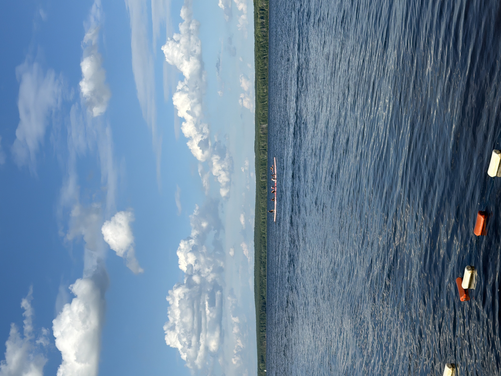 A wide shot captures a serene summer day on the Kama River in Chaikovsky, Russia. The water, a deep blue, is gently rippled, reflecting the bright, partly cloudy sky above. In the distance, a lush green forest forms a dense treeline along the horizon. Several people are visible on a long, pink boat, rowing in unison across the river. In the foreground, orange and white buoys float on the water's surface, marking a designated area. The scene conveys a sense of peaceful recreation and natural beauty.
