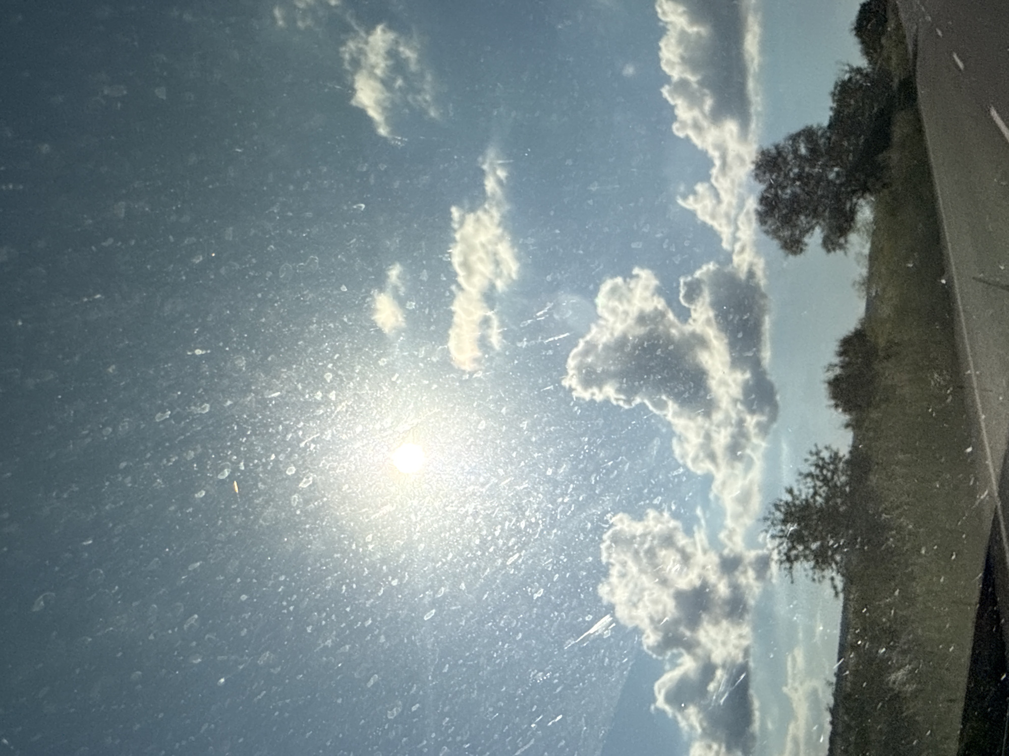 The image depicts a view through a dirty windshield, looking out at a sunny sky with scattered clouds. The sun is bright and positioned in the upper left quadrant of the frame, casting a glare. The sky is a clear blue, transitioning to lighter shades near the horizon. Several fluffy white clouds are visible, with some darker, more textured clouds in the middle ground. Below the sky, a landscape of green grass and trees stretches across the horizon. A road, possibly asphalt, runs diagonally from the lower right to the lower left of the frame, with white lines marking the lanes. The overall impression is that of a scenic drive through a rural area. The specific location in the city of Yabalchevo, Bulgaria, is not discernible from the visual information provided.