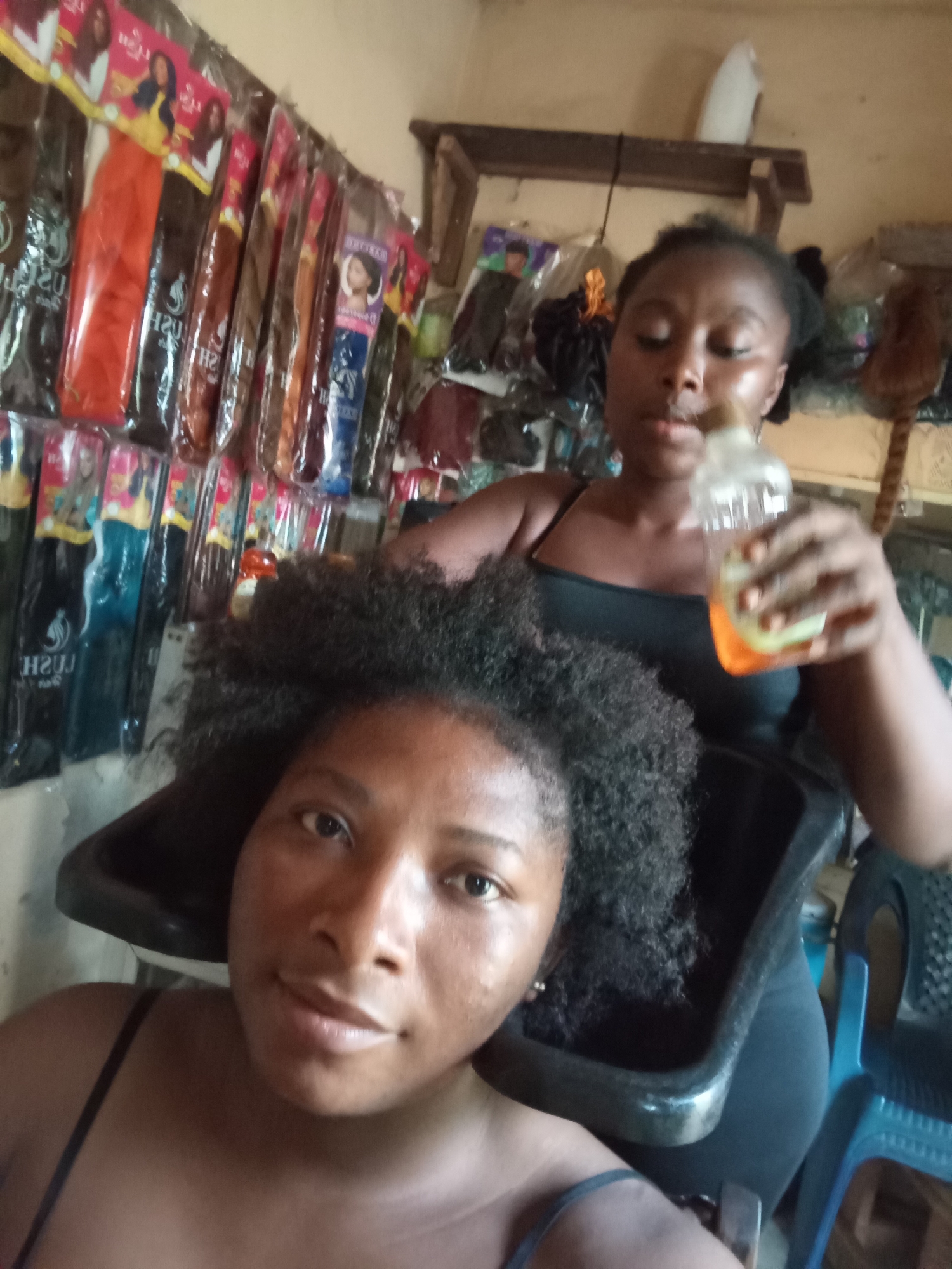 The photograph appears to be taken inside a small hair salon in Nkpor, Nigeria. The scene shows two women. In the foreground, a woman sits facing the camera, presumably a client. Her hair is afro-textured and appears to be dry. Her head is resting in a black plastic hair washing basin. Behind her stands another woman, likely a hair stylist, holding a clear plastic bottle with an orange liquid inside. The stylist is wearing a black tank top. On the wall behind the women, there is a display of packaged hair extensions in various colors. In the background, there are some additional hair products and salon equipment. A blue plastic chair is also visible on the right side of the image. The lighting is somewhat dim and appears to be a combination of natural light and indoor lighting. The salon has a simple and functional decor.
