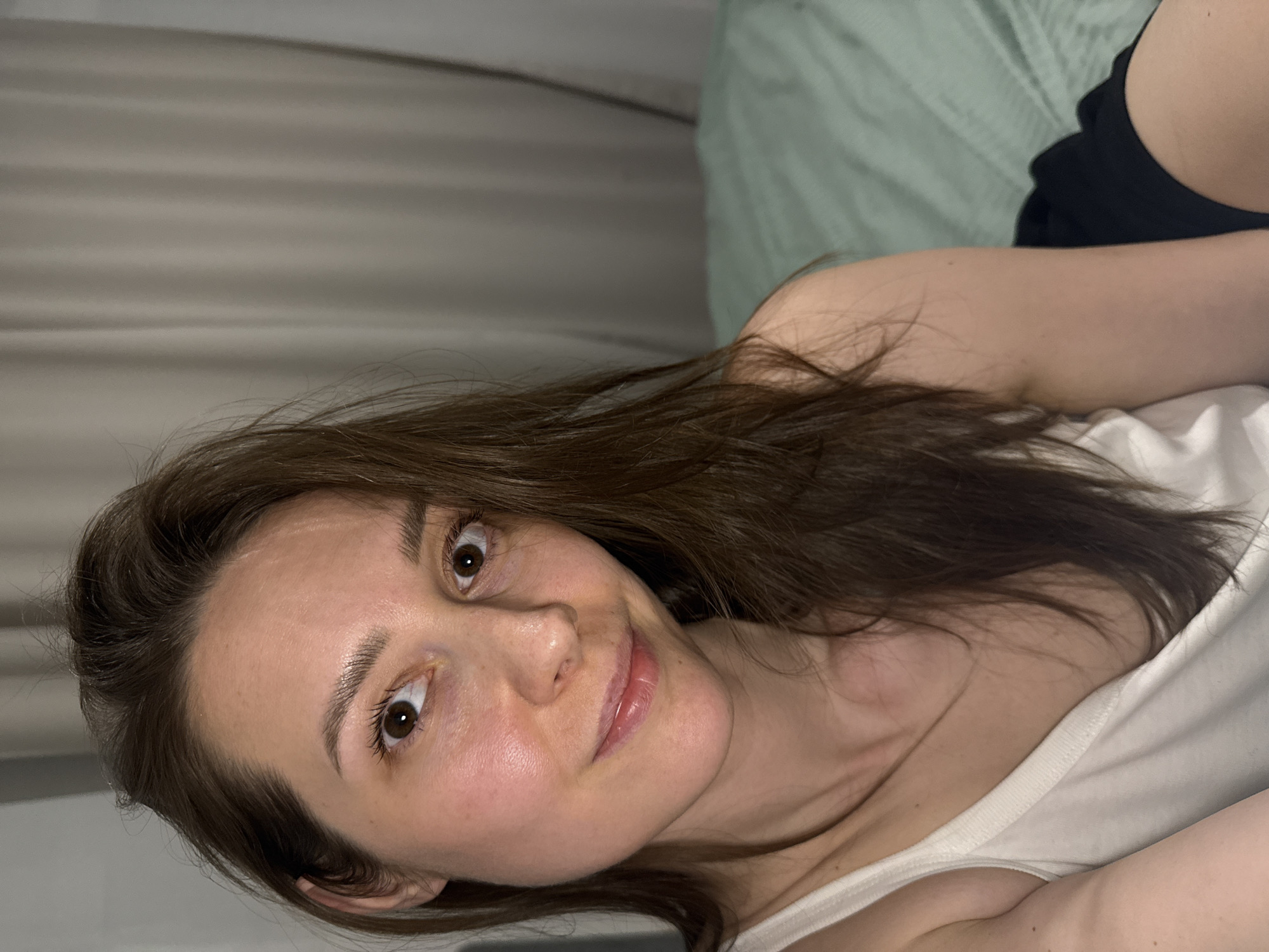 A young adult female with light skin, long brown hair, and dark brown eyes is depicted in an indoor setting, likely a bedroom, within the city of Chaikovsky, Russia. She is facing forward, looking directly at the camera with a slight, closed-lip smile. Her hair is parted and falls over her shoulders. She is wearing a white tank top. The background on the left features light-colored curtains with vertical folds. On the right, a bed with a light green striped duvet or sheet is visible. A dark fabric, possibly part of her attire, is seen near her right hip. Her left arm is partially visible in the lower-left foreground. The lighting is soft, illuminating her face.