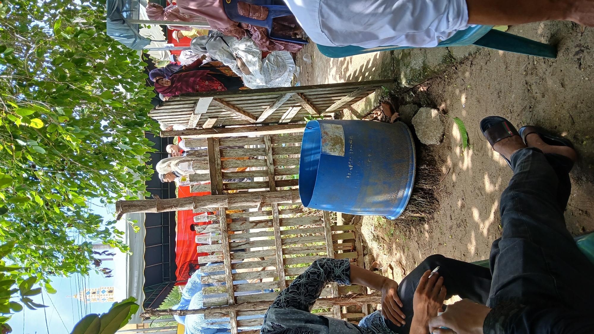 The image shows an outdoor scene in Lhokseumawe, Indonesia. People are seated near a wooden fence and a large blue plastic drum. Some individuals are wearing traditional Muslim attire. One person is holding a cigarette. Overhead, there are leafy trees and glimpses of a structure, possibly a minaret, suggesting a location near a mosque or religious building. Clothes are also seen hanging overhead. The ground is dirt.