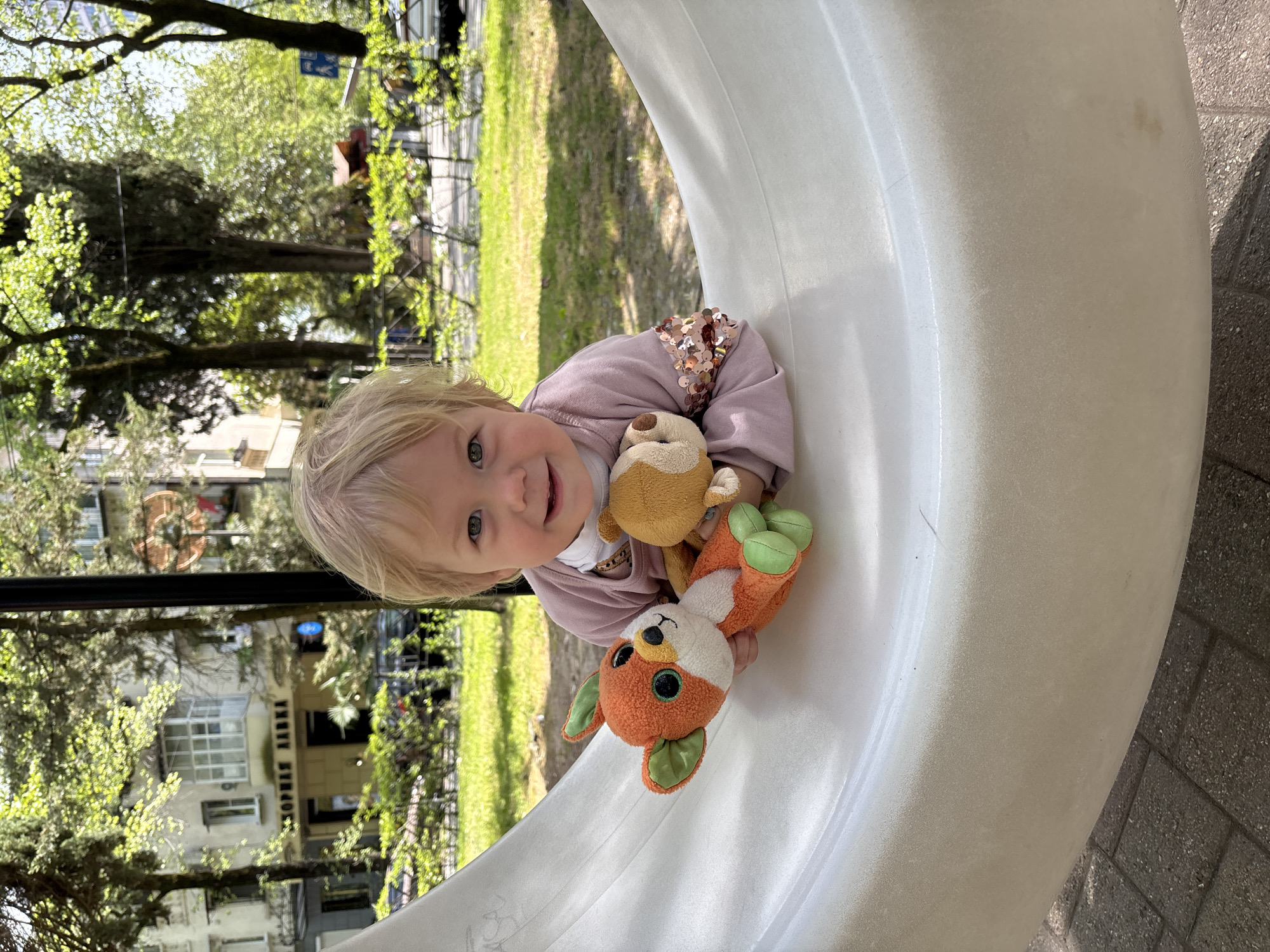 A young, blond-haired child wearing a light purple long-sleeved shirt with sequins is sitting inside a large, curved, white structure, holding two stuffed animals. The child is smiling and looking directly at the camera. The stuffed animals include a small brown creature and an orange fox with green ears.
Behind the child, green trees and grass are visible, suggesting a park-like setting. A building with a sign "МОРЕМА АДКА" is also visible in the background. Given the information provided, the location is likely Sochi, Russia. The surface of the white structure the child is in shows signs of wear and slight discoloration.