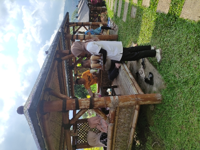The image depicts a group of people sitting and standing under a bamboo gazebo in Kecamatan Pujon, Indonesia. The gazebo is made of bamboo and has a thatched roof. The people are sitting and standing on a wooden platform that is surrounded by green grass. There are several stone paths leading away from the gazebo. The people are dressed in casual clothing. One woman is holding a tray with multiple glasses of beverages. The people are interacting with each other and appear to be enjoying their time together. There are a few sandals on the ground, suggesting people have recently removed footwear. The sky is blue and cloudy, and there is a green hillside in the distance. The coordinates provided are: -7.854, 112.453.