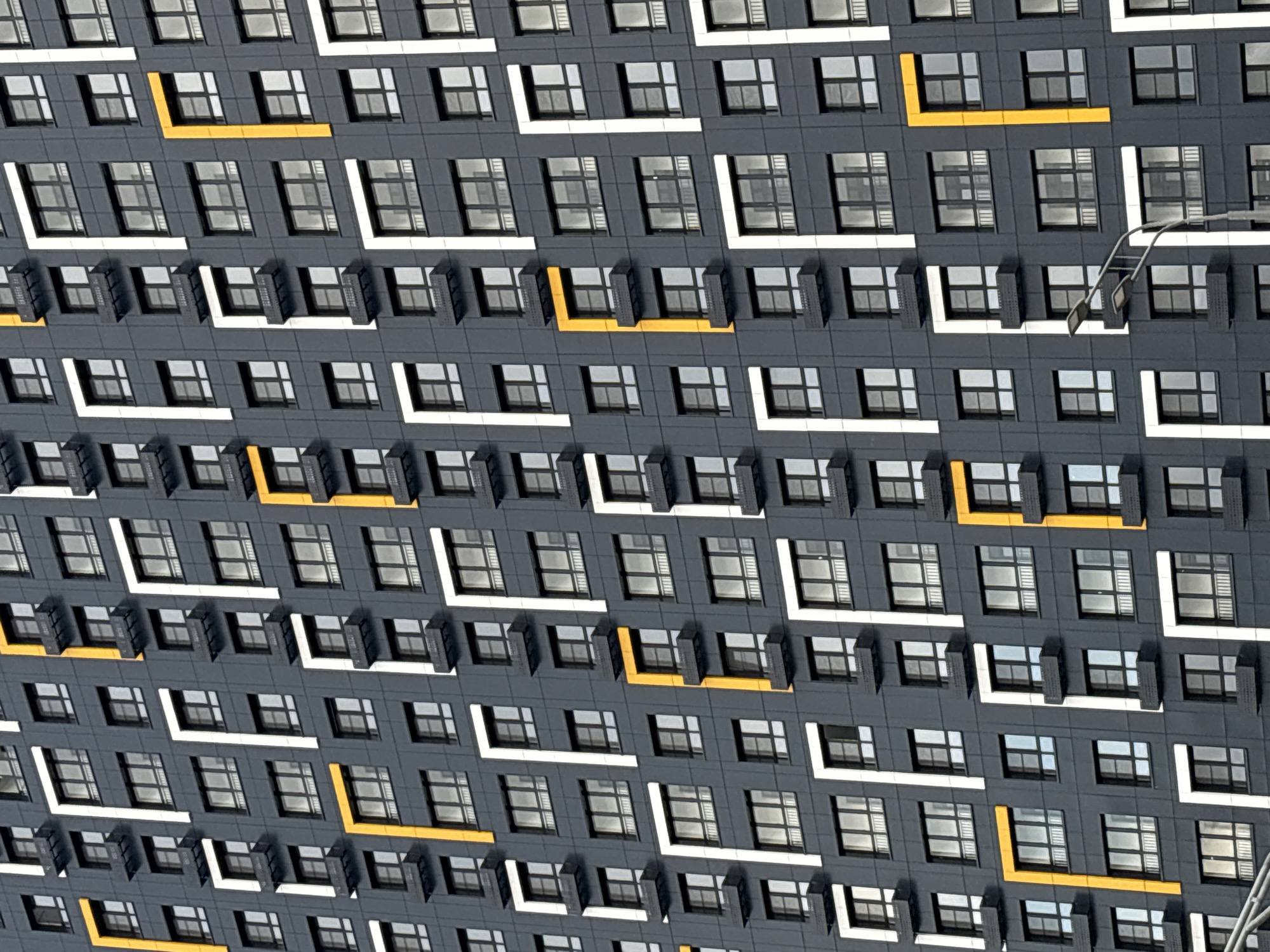 This is a full-frame, eye-level, medium shot of the facade of a modern residential building in Moscow, Russia. The building features a repeating pattern of dark gray panels, with alternating sections of windows and decorative white and yellow geometric accents. Each window unit is recessed and appears to have a small balcony area with a dark railing. The geometric accents form L-shapes and vertical stripes, creating a visually striking, almost pixelated effect. The lighting suggests it is daytime, with the sun casting subtle shadows on the building's surfaces. No people are visible in the image. The scene is an urban architectural study focusing on the design and material of the building.