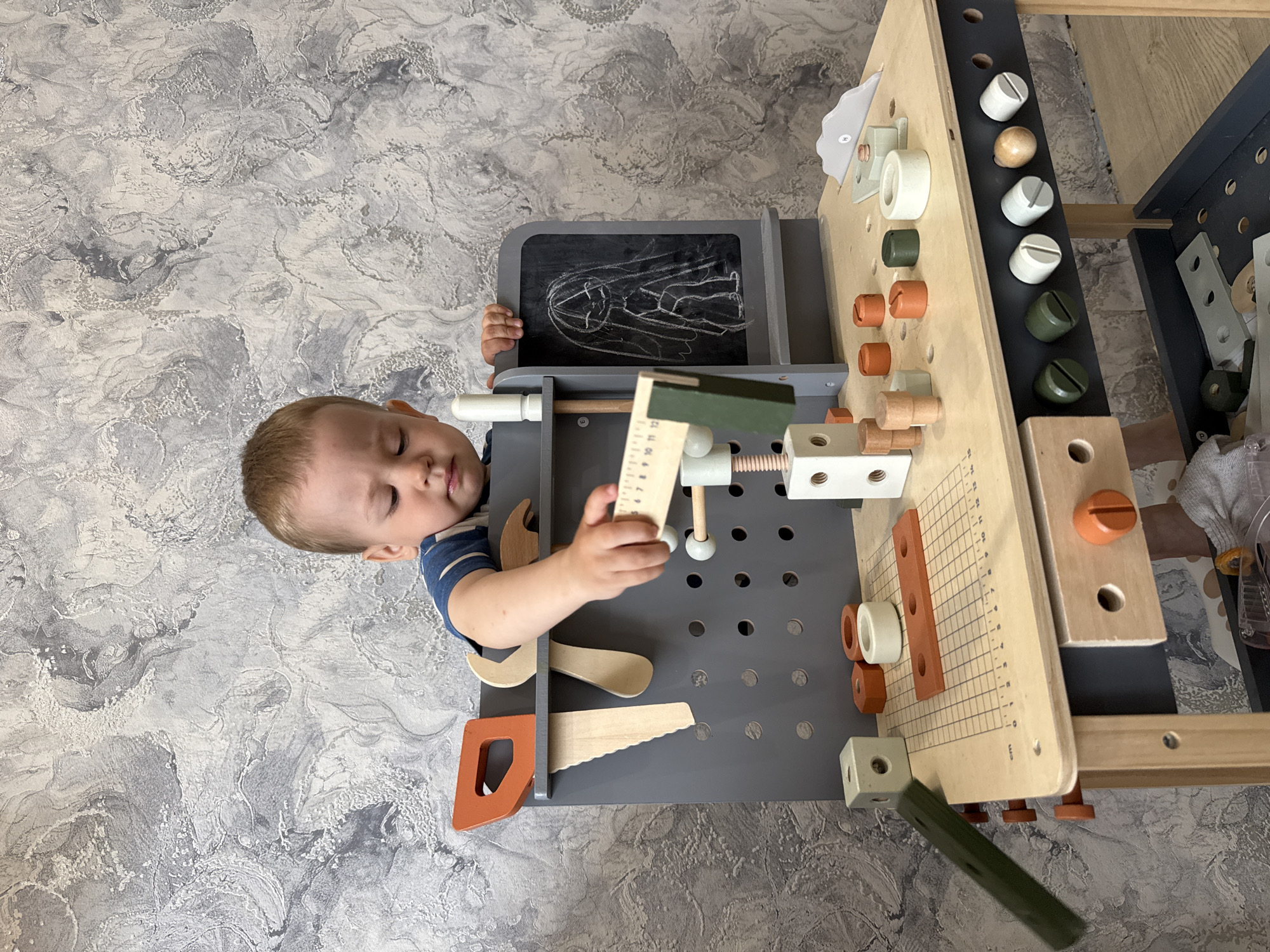 The image shows a young child, likely under the age of 3, lying on his stomach on a gray, patterned surface resembling marble. The child is playing with a wooden toy workbench, which is equipped with various tools and accessories like screws, measuring stick and wooden pieces. The workbench is located on the surface with the child. The background consists of the same gray, patterned surface. The location could be Rusanovo, Russia.