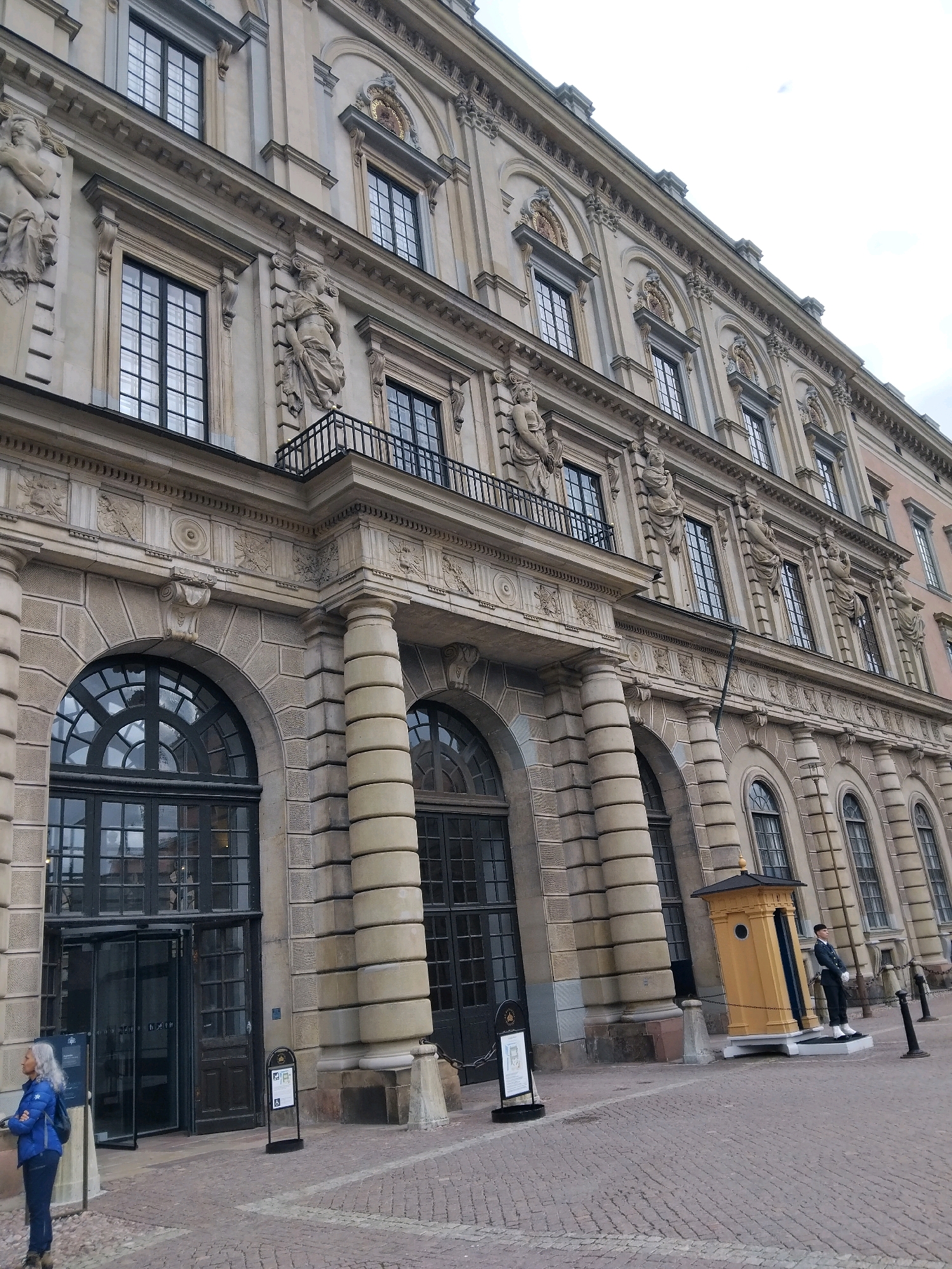 This image depicts the Royal Palace in Stockholm, Sweden, captured from a low angle looking upwards at the facade. The ornate sandstone building features multiple stories with numerous windows, decorative sculptures, and classical architectural elements like columns and arches. In the foreground, a cobblestone plaza is visible, with a yellow guardhouse and a uniformed guard standing to the right. To the left, a woman in a blue jacket stands near an entrance with glass doors. The sky is overcast.