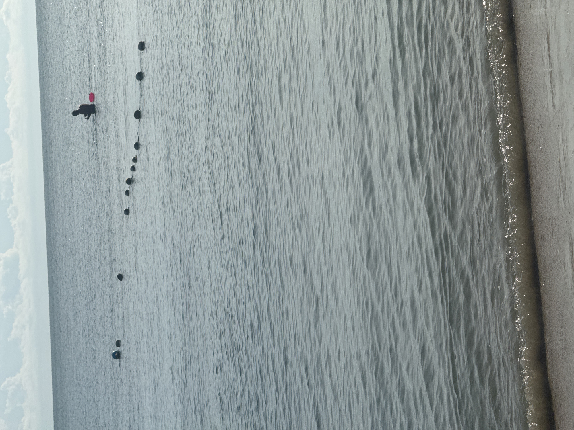 A person stands in the ocean with a red object nearby, positioned behind a line of buoys. The scene is a calm body of water with a sandy shoreline in the foreground. The sky above is partly cloudy. The location is unknown.