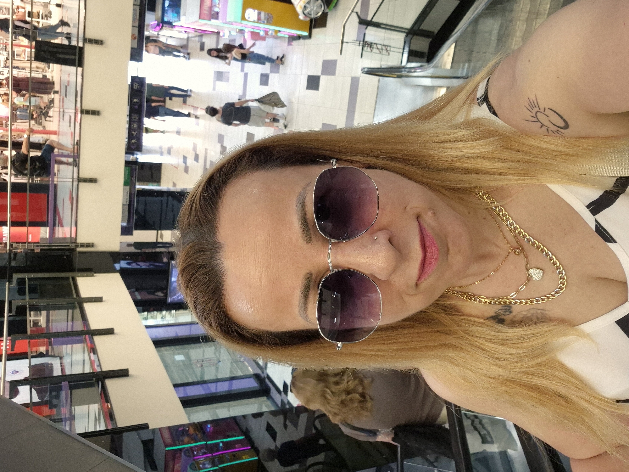 A woman with blonde hair and sunglasses takes a selfie in a shopping mall. She is wearing a white and black top and multiple gold necklaces. She has a tattoo on her right arm and a piercing on her nose. In the background, other shoppers are visible on different levels of the mall, some on escalators. The mall appears to be multi-storied with shops and displays visible.